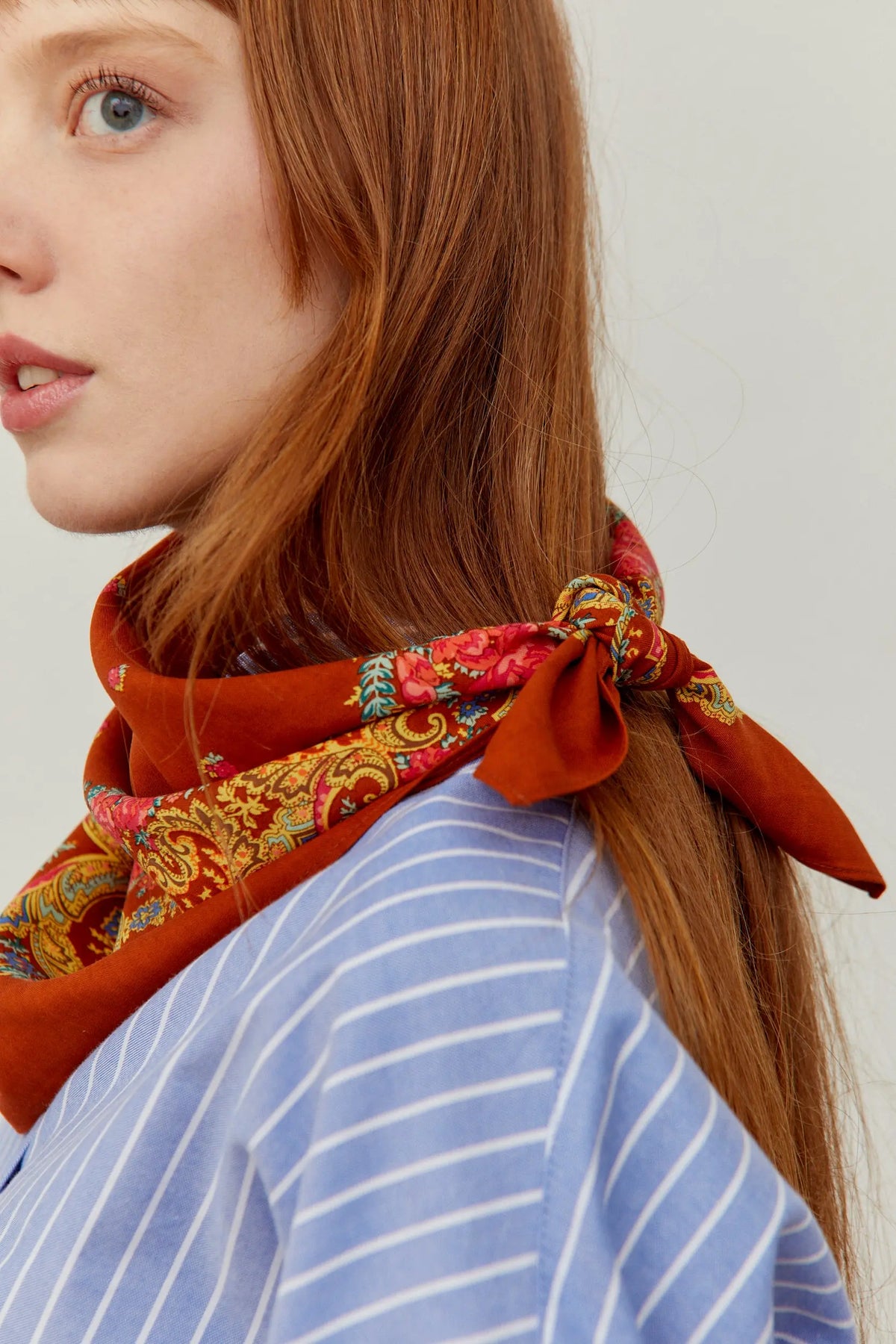 Rust-colored scarf with a traditional floral chain print, tied around the neck, worn with a blue striped shirt.