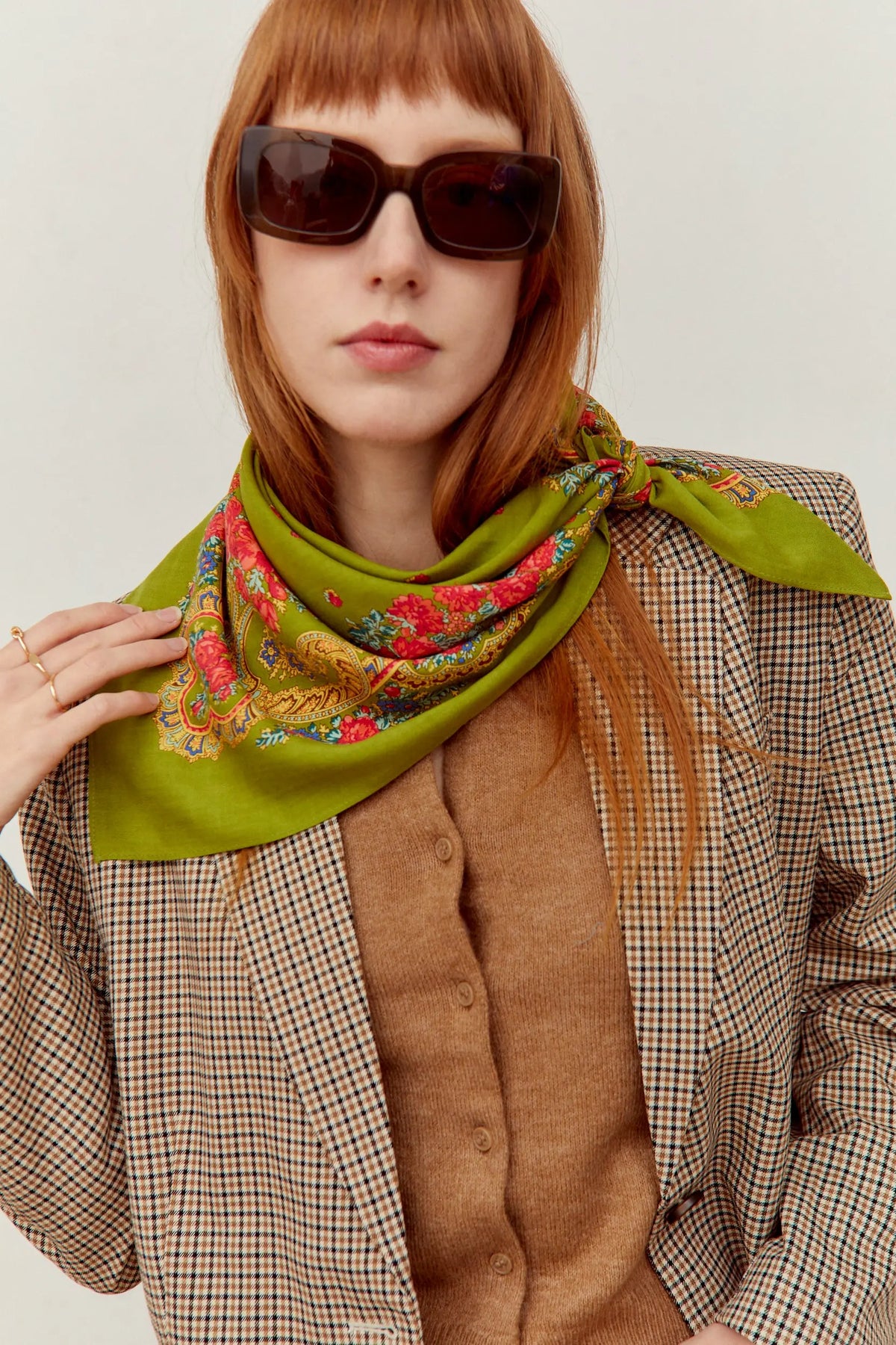Pañuelo verde oliva con estampado floral y de cadenas, estilo clásico, anudado al cuello sobre chaqueta a cuadros.