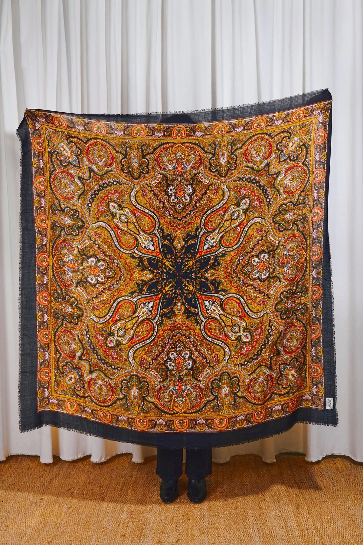 Pañuelo de lana negro con estampado tipo cachemira en tonos naranja, amarillo y blanco, de forma cuadrada y borde desflecado.