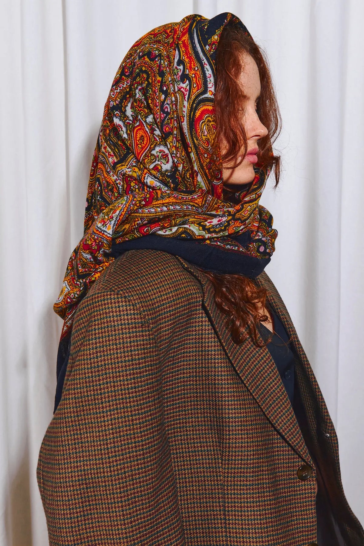 Pañuelo de lana negro con estampado paisley multicolor y vibrante sobre la cabeza de una mujer con blazer de cuadros.