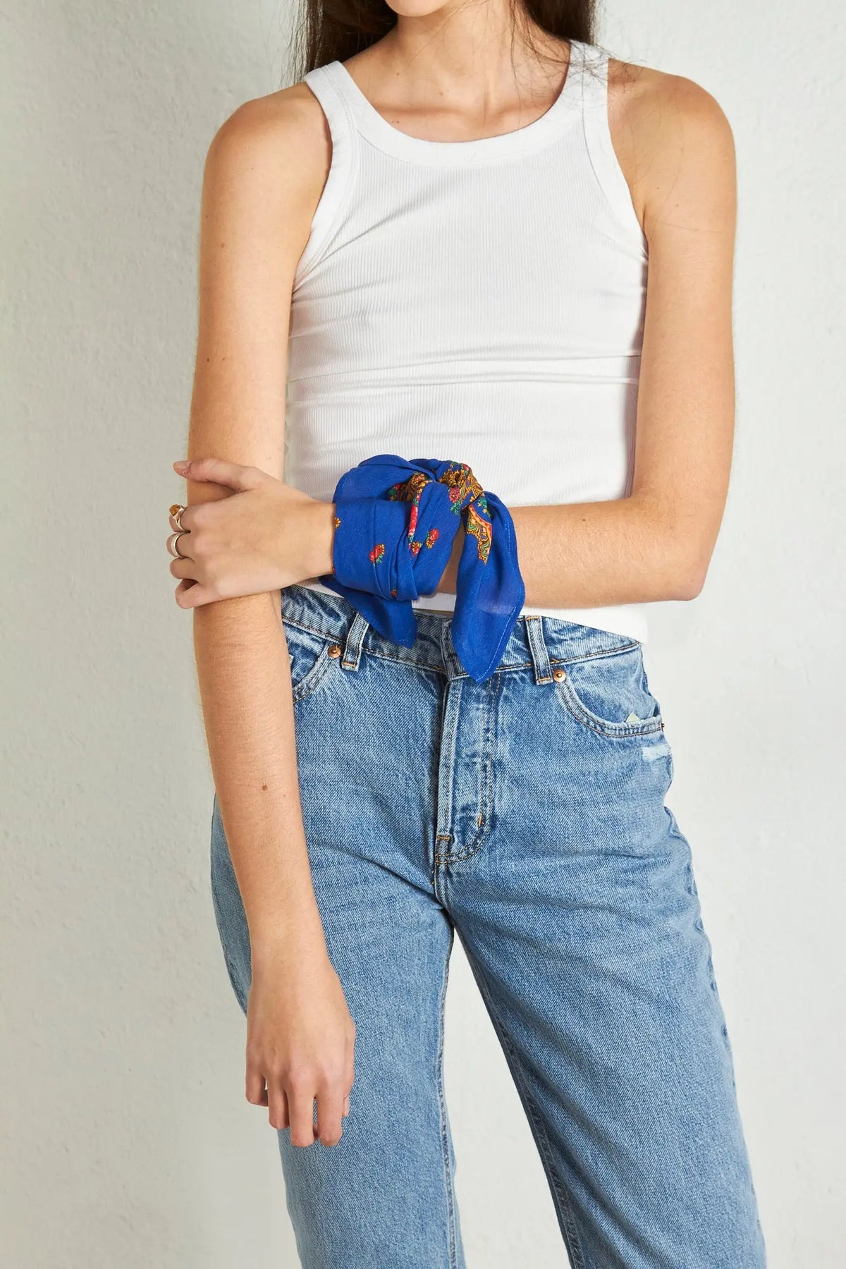 A blue square scarf with a gold chain and floral pattern tied around a woman’s wrist, worn with a white ribbed tank.