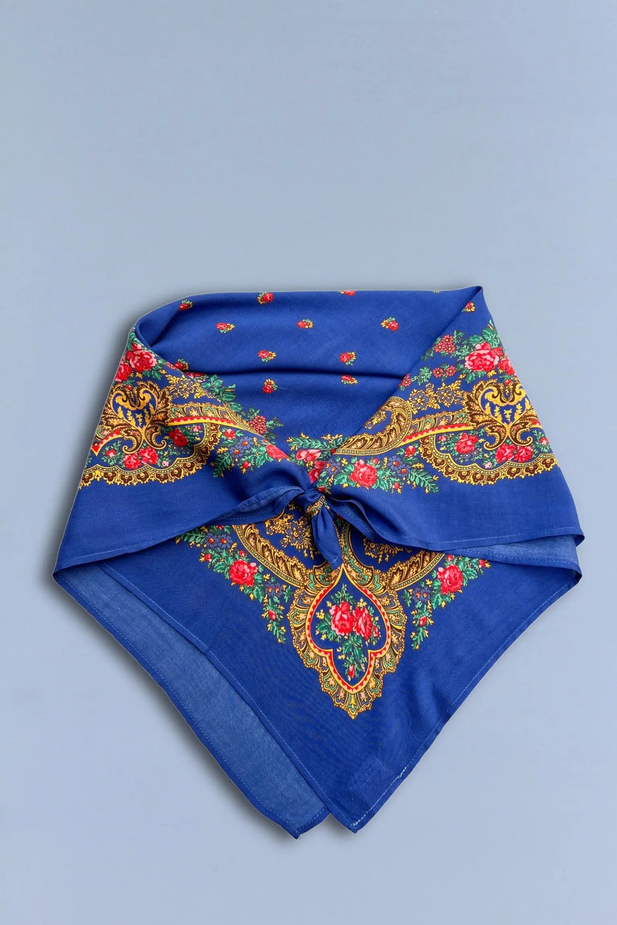 Pañuelo azul anudado con estampado floral tradicional, detalles de cadenas doradas y flores rojas sobre fondo de tela.