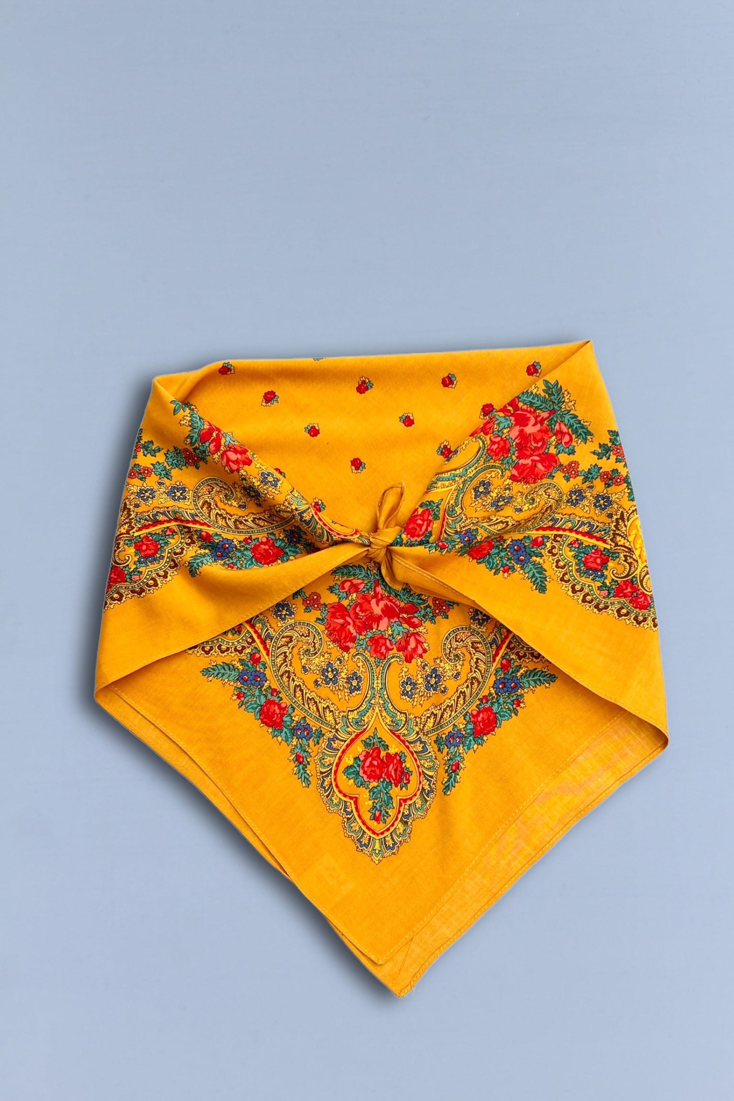 Pañuelo ocre de estilo tradicional con estampado de flores rojas, detalles en azul y cadenas sobre fondo liso.