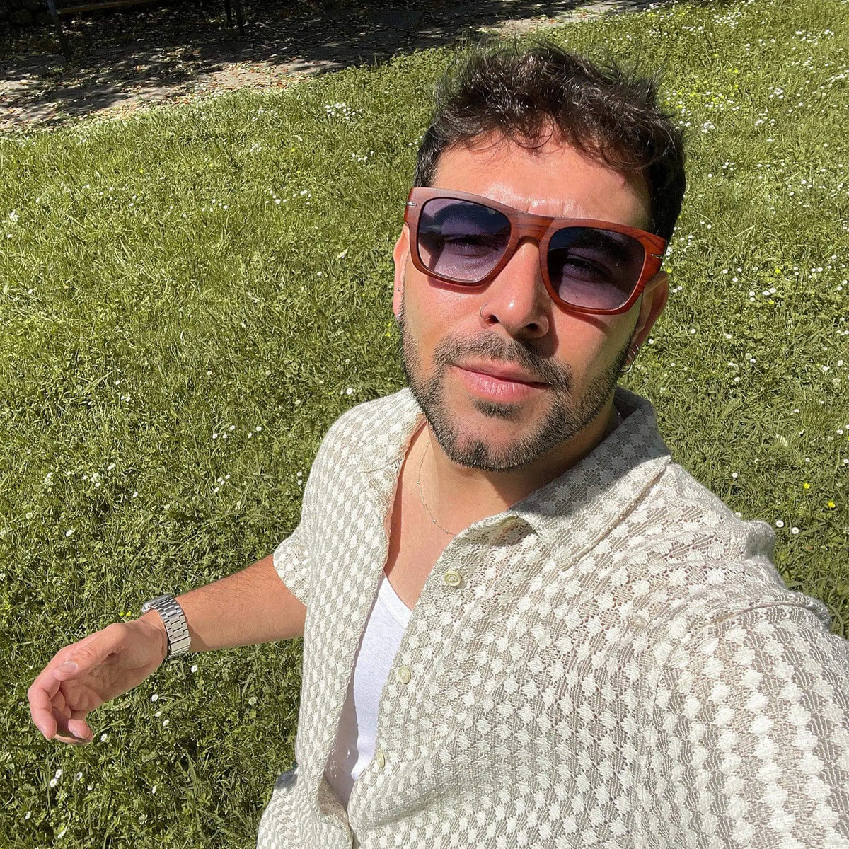 Hombre con gafas de sol cuadradas de madera con vetas y lentes azules en un jardín soleado.