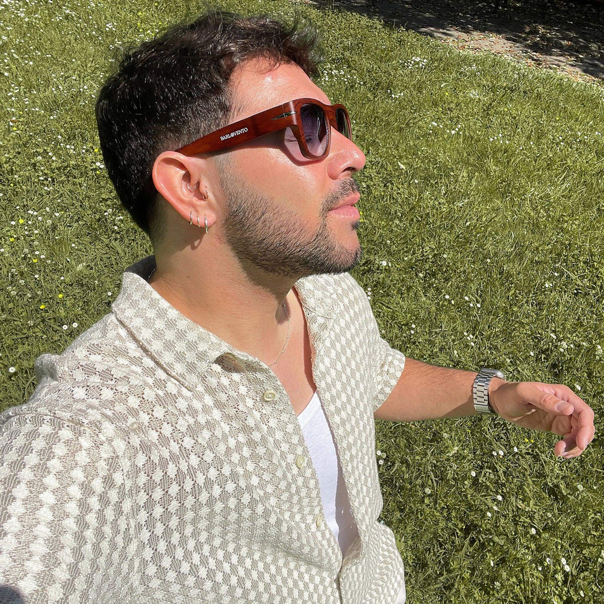 Hombre con gafas de sol cuadradas efecto madera y lentes azules en un jardín, vistiendo camisa texturizada clara.