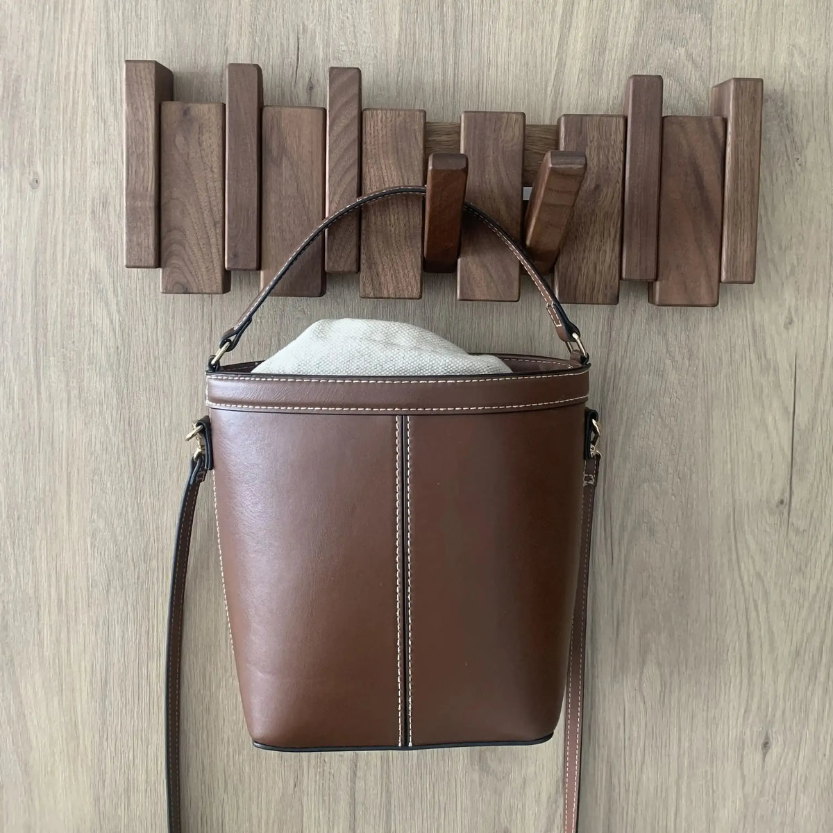 Perchero de pared de madera oscura con piezas plegables de estilo escandinavo, sosteniendo un bolso de cuero marrón.