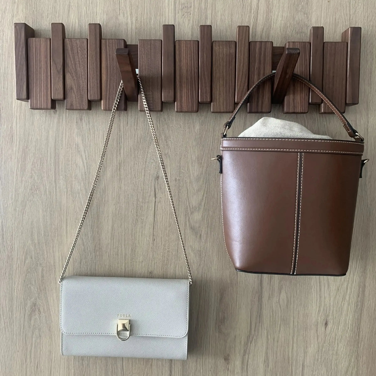 Perchero de pared plegable en madera oscura de estilo escandinavo con bolsos colgados, sobre una pared de madera clara.