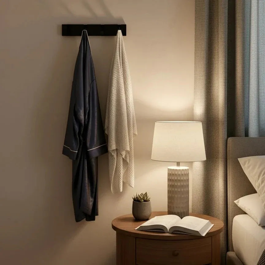 Perchero de pared moderno en metal negro con batas colgadas sobre una mesita de noche en un dormitorio cálido.