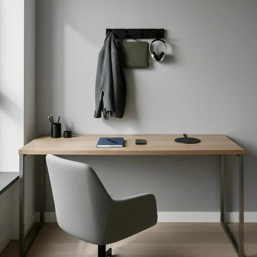 Perchero de pared moderno en metal negro instalado sobre una pared gris, con gancho para ropa, bolso y auriculares.