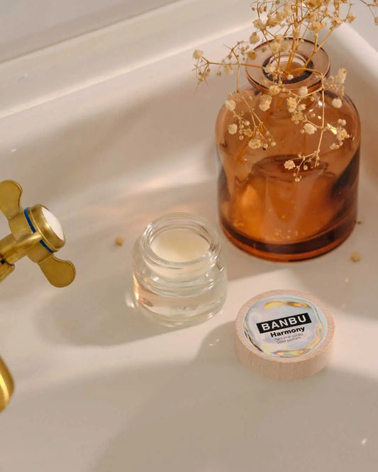 Perfume sólido Banbu Harmony en tarro de cristal, junto a un jarrón color ámbar y flores secas en un lavabo blanco.