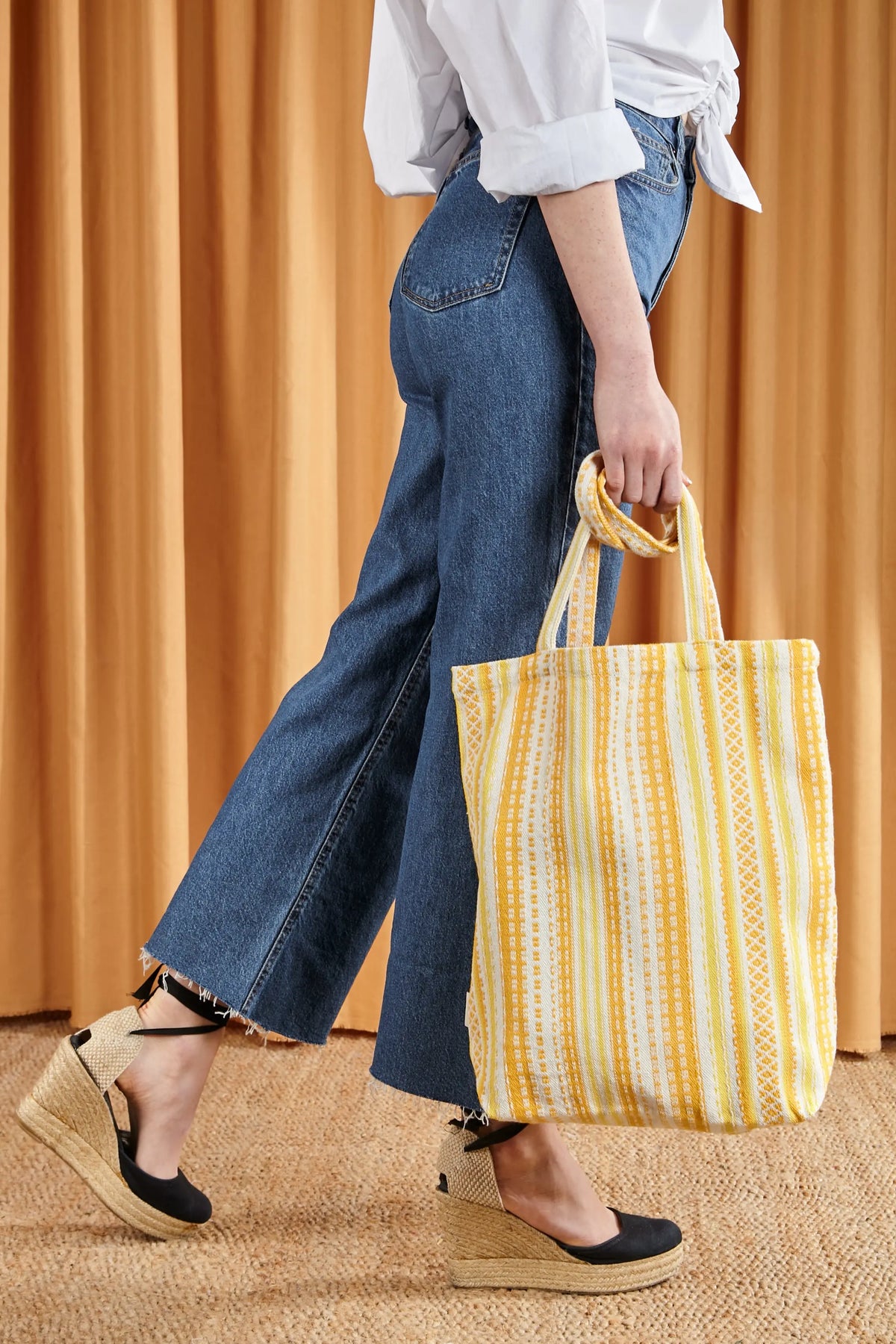 Tote bag de tela con estampado a rayas en tonos amarillos y estilo artesanal, sostenida por una persona en jean.