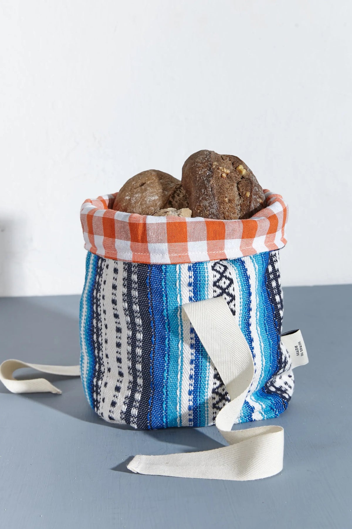 Panera de tela con estampado alpujarreño en tonos azules, interior de cuadros naranjas y asas de lona cruda.