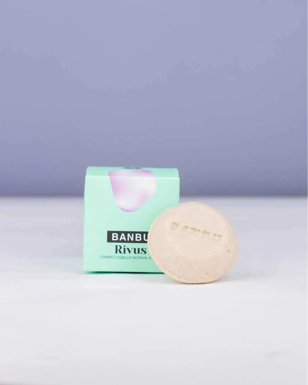 Champú sólido redondo y beige de Banbu junto a su caja de cartón verde claro sobre una superficie blanca.