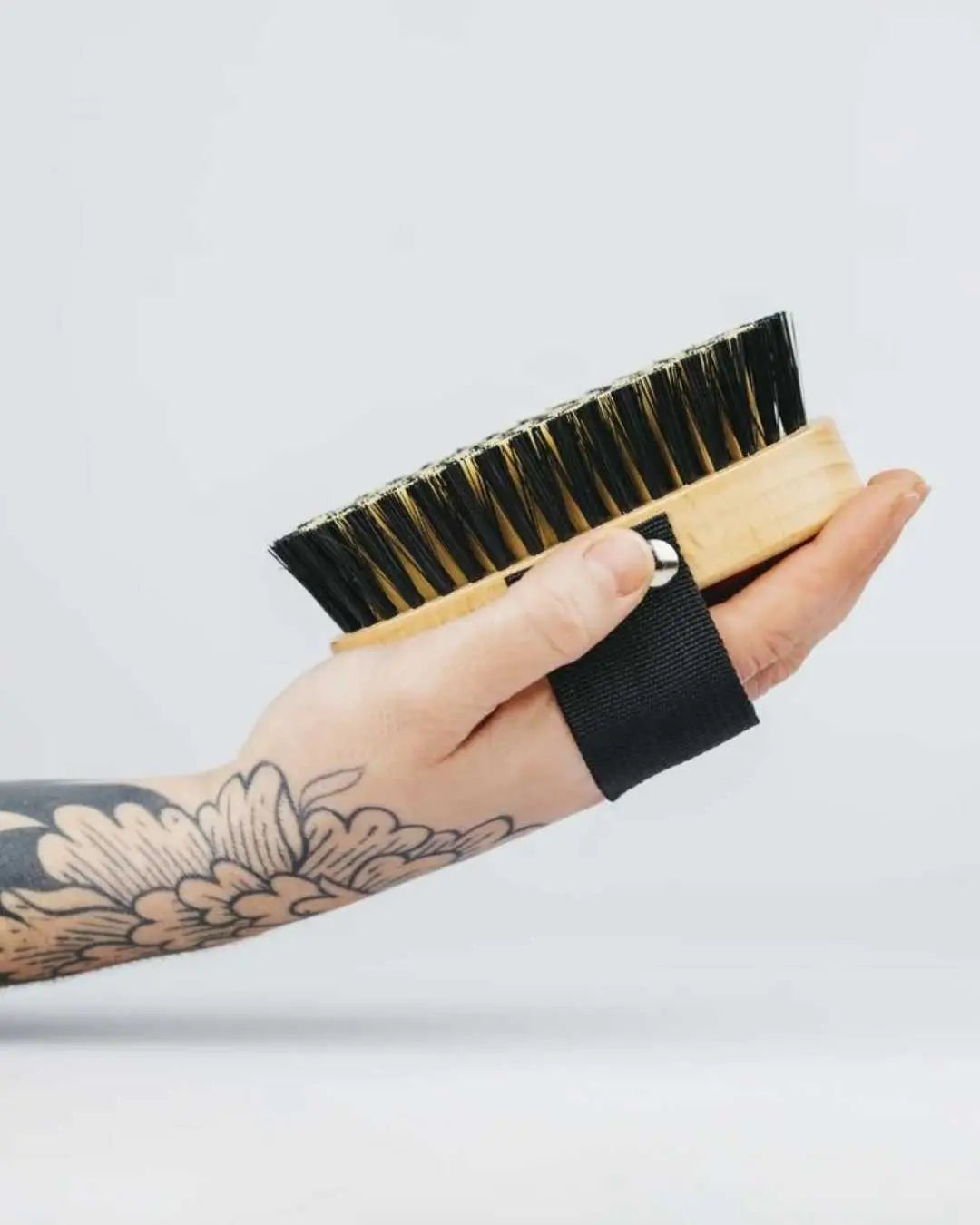 Mano con tatuaje sostiene un cepillo Banbu de madera clara con cerdas negras y cinta de sujeción negra.