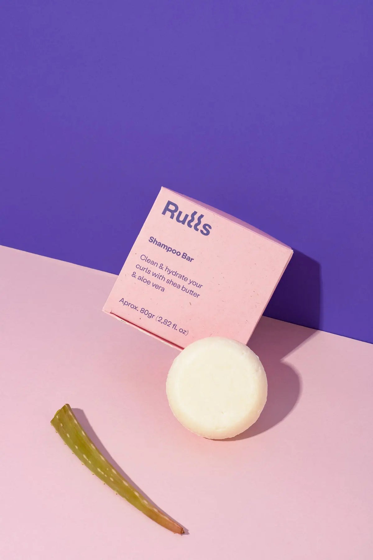 Barra de champú blanca con envoltura rosa, contiene manteca de karité y aloe vera, para cabello rizado.