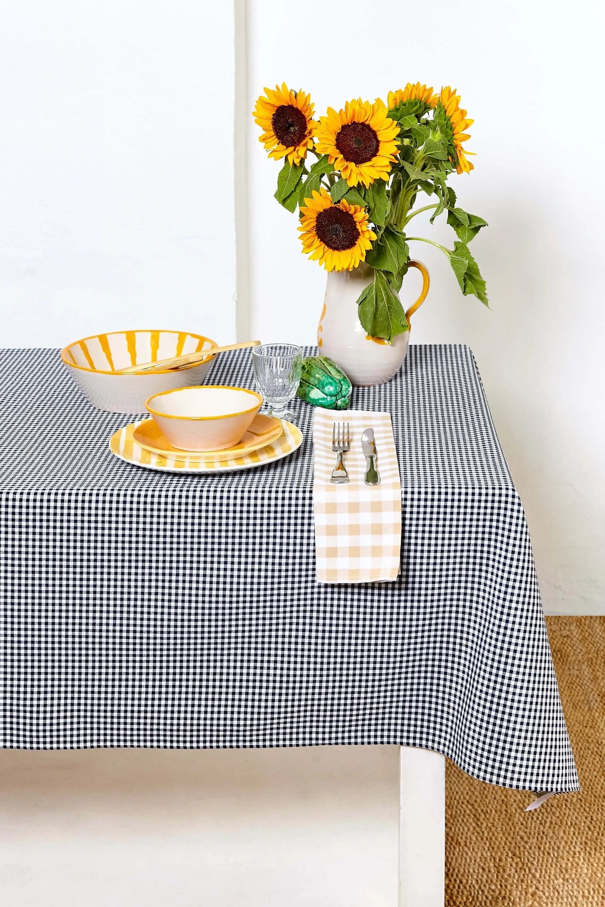 Mantel de tela con estampado de cuadros vichy negros y blancos sobre una mesa con vajilla y un jarrón con girasoles.
