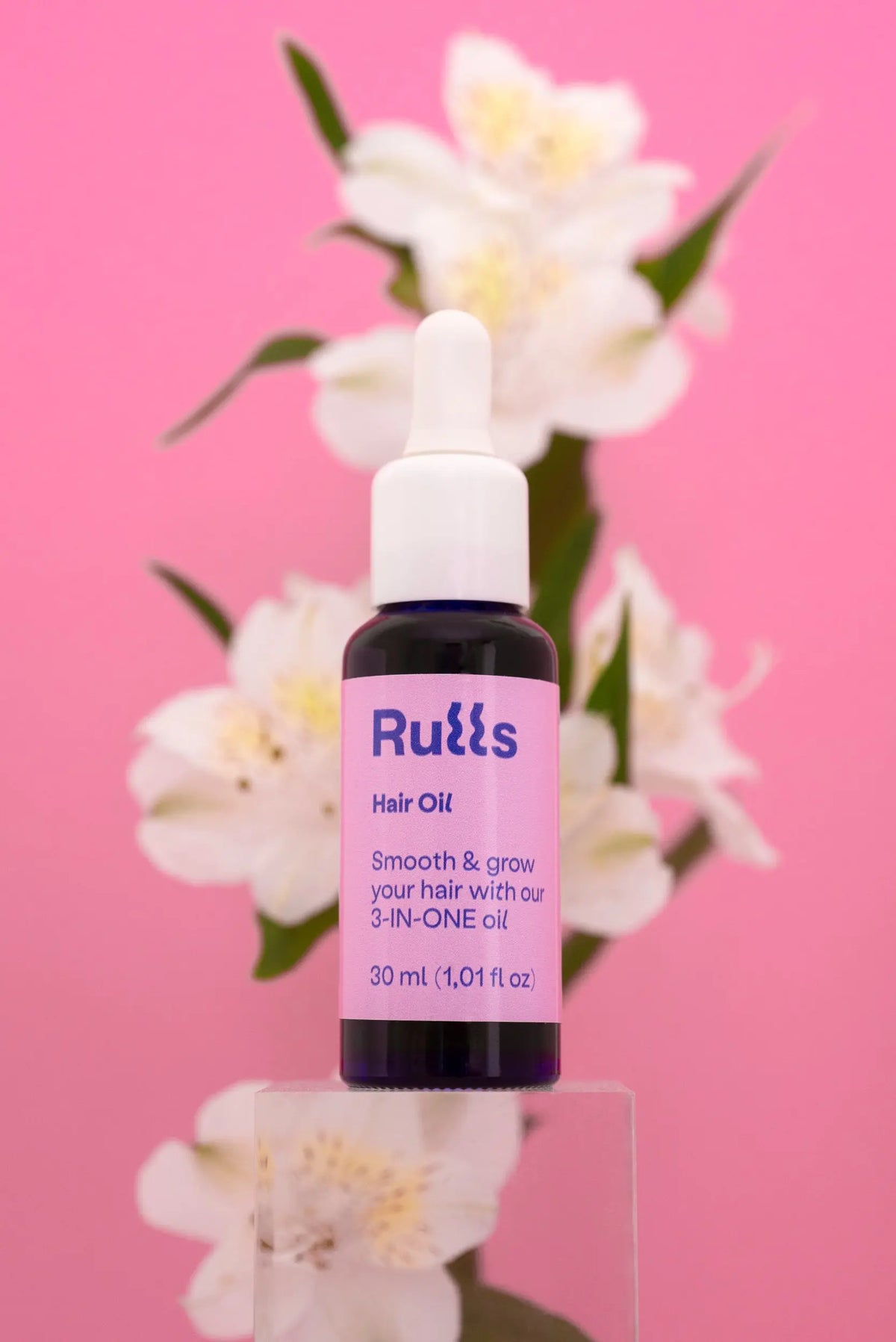 Aceite para el cabello Rulls 30 ml, frasco negro con etiqueta morada, fondo rosa y flores blancas.