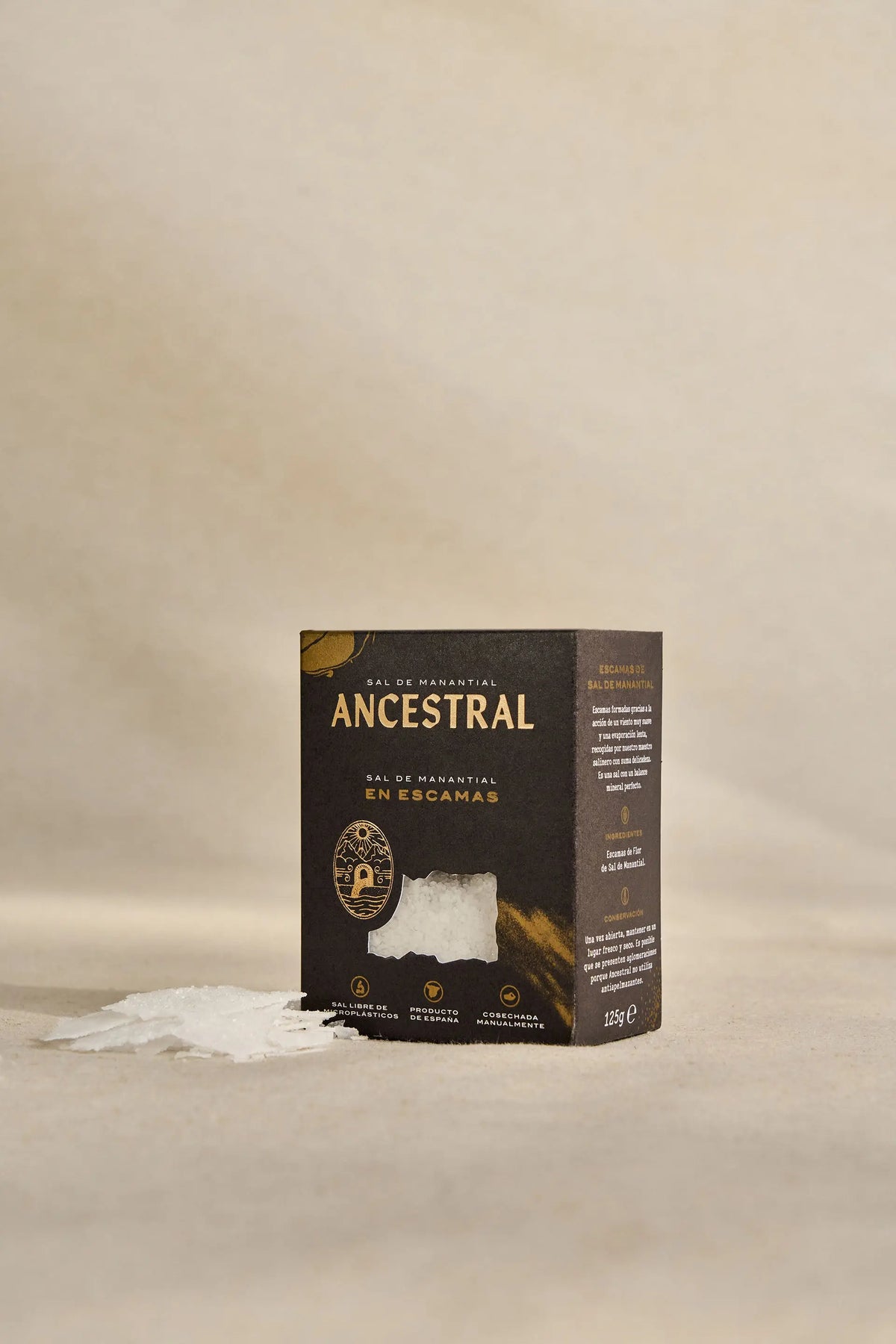 Black box of gourmet spring salt flakes with a clear window, placed next to a small pile of salt flakes on beige.