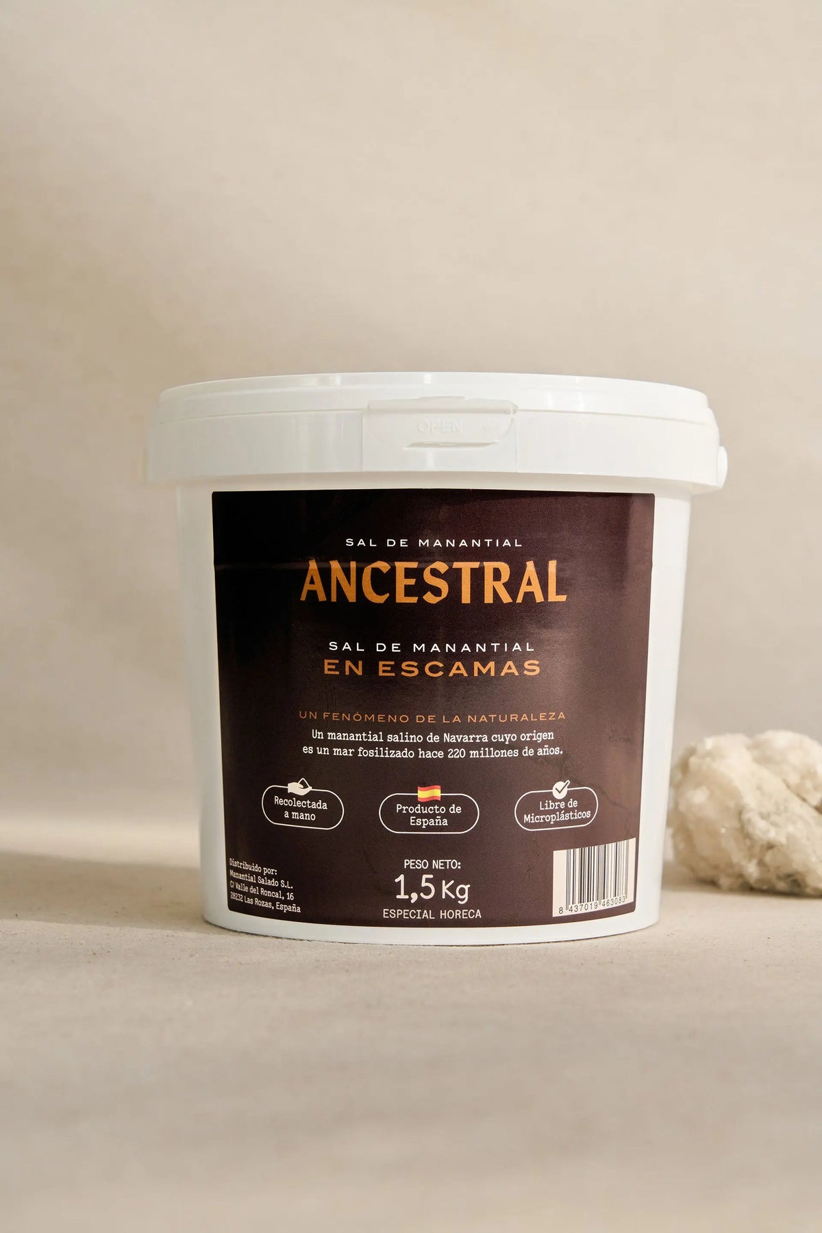 White plastic bucket of gourmet spring salt flakes with a dark brown label and a rough salt rock beside it.