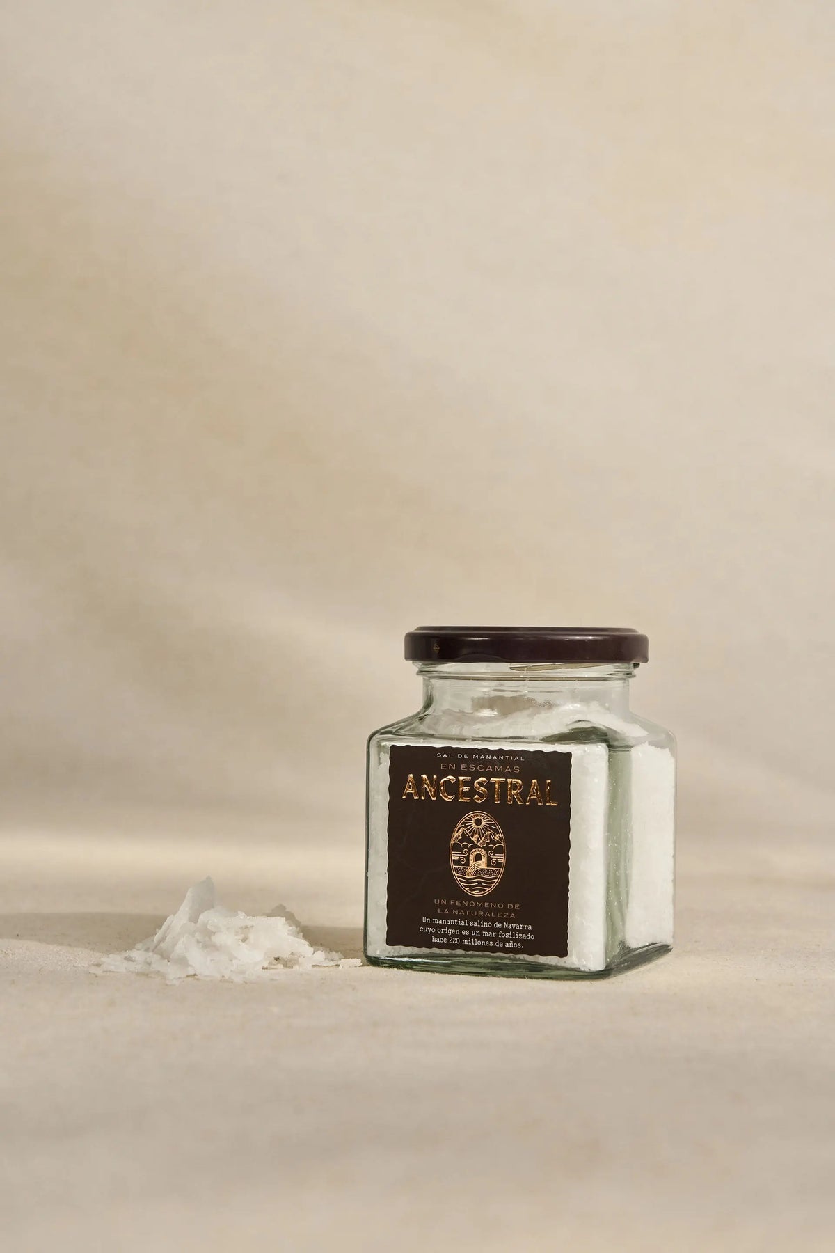 Glass jar of gourmet salt flakes with a dark label, featuring a small pile of salt beside it on a beige background.