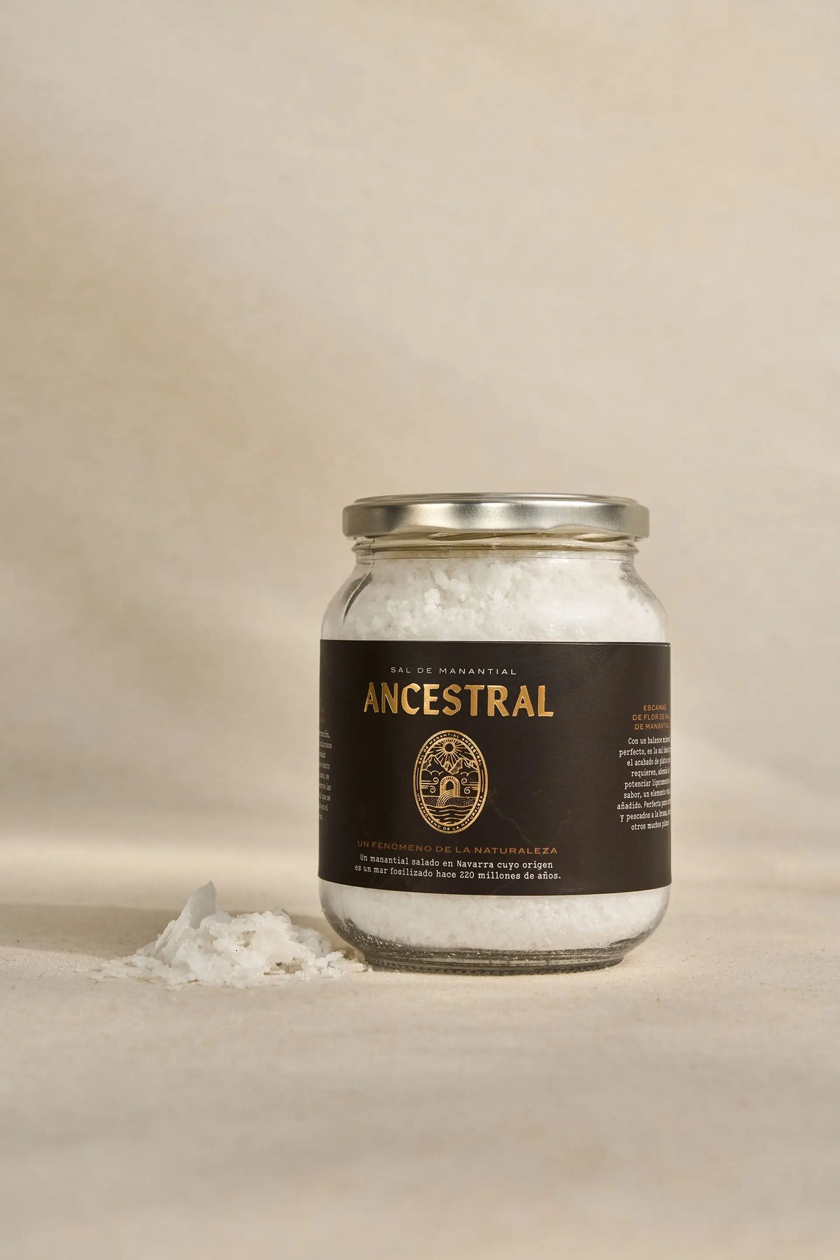 Glass jar of white spring salt flakes with a dark, gold-lettered Ancestral label on a beige, textured background.