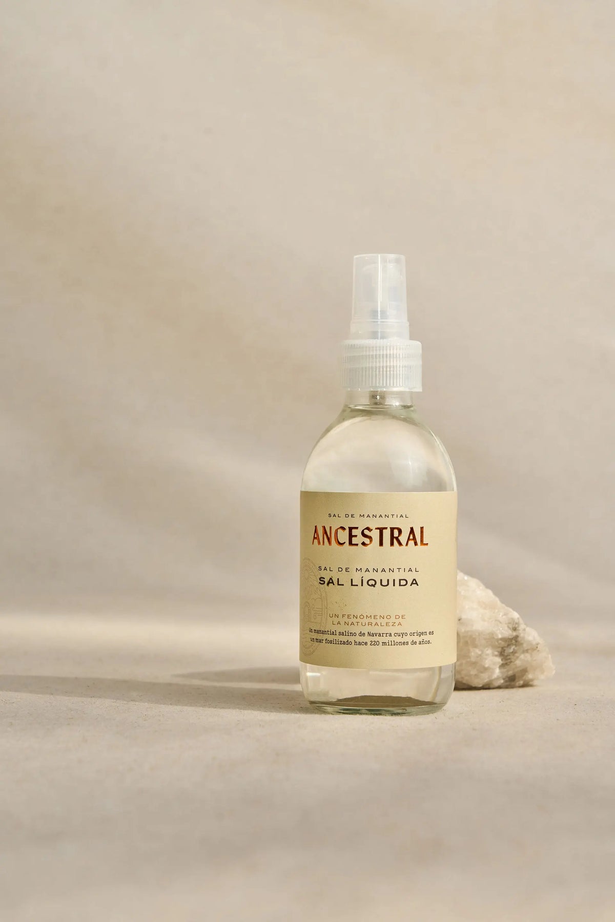 Clear glass spray bottle of liquid salt with a beige label, positioned next to a piece of raw mineral on a tan surface.