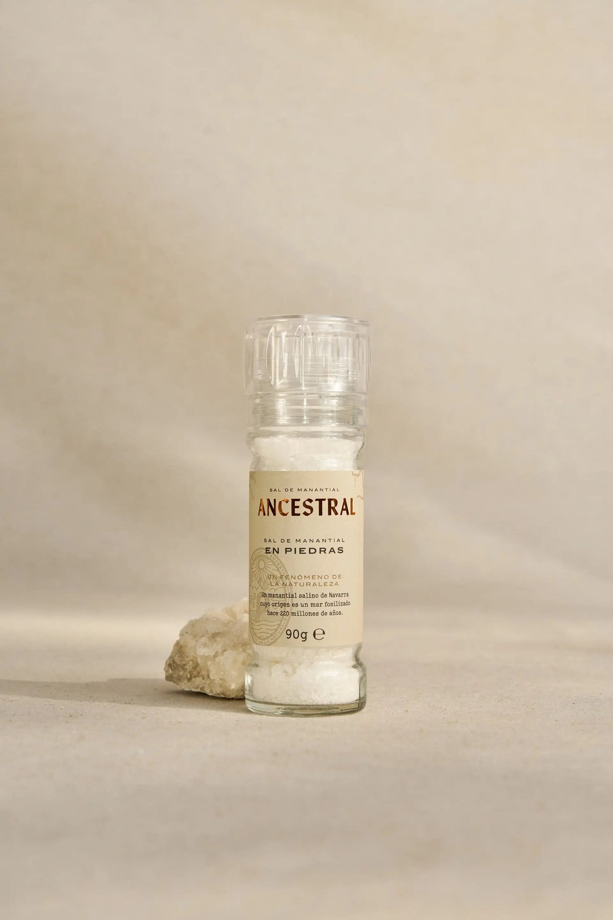 Glass salt mill filled with natural rock salt crystals, placed next to a raw salt stone on a beige background.