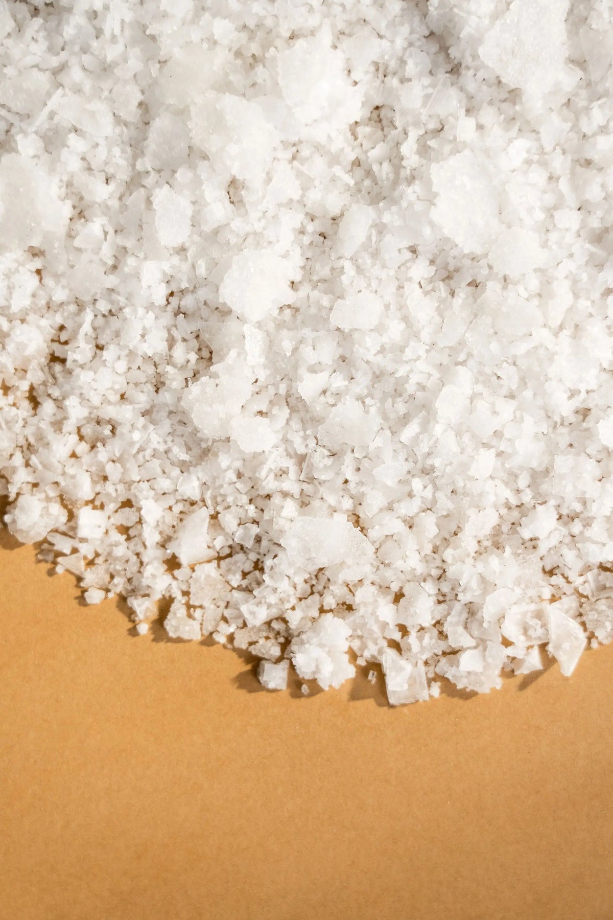 White, irregular, flaky spring salt crystals scattered on a smooth, warm tan-colored surface.