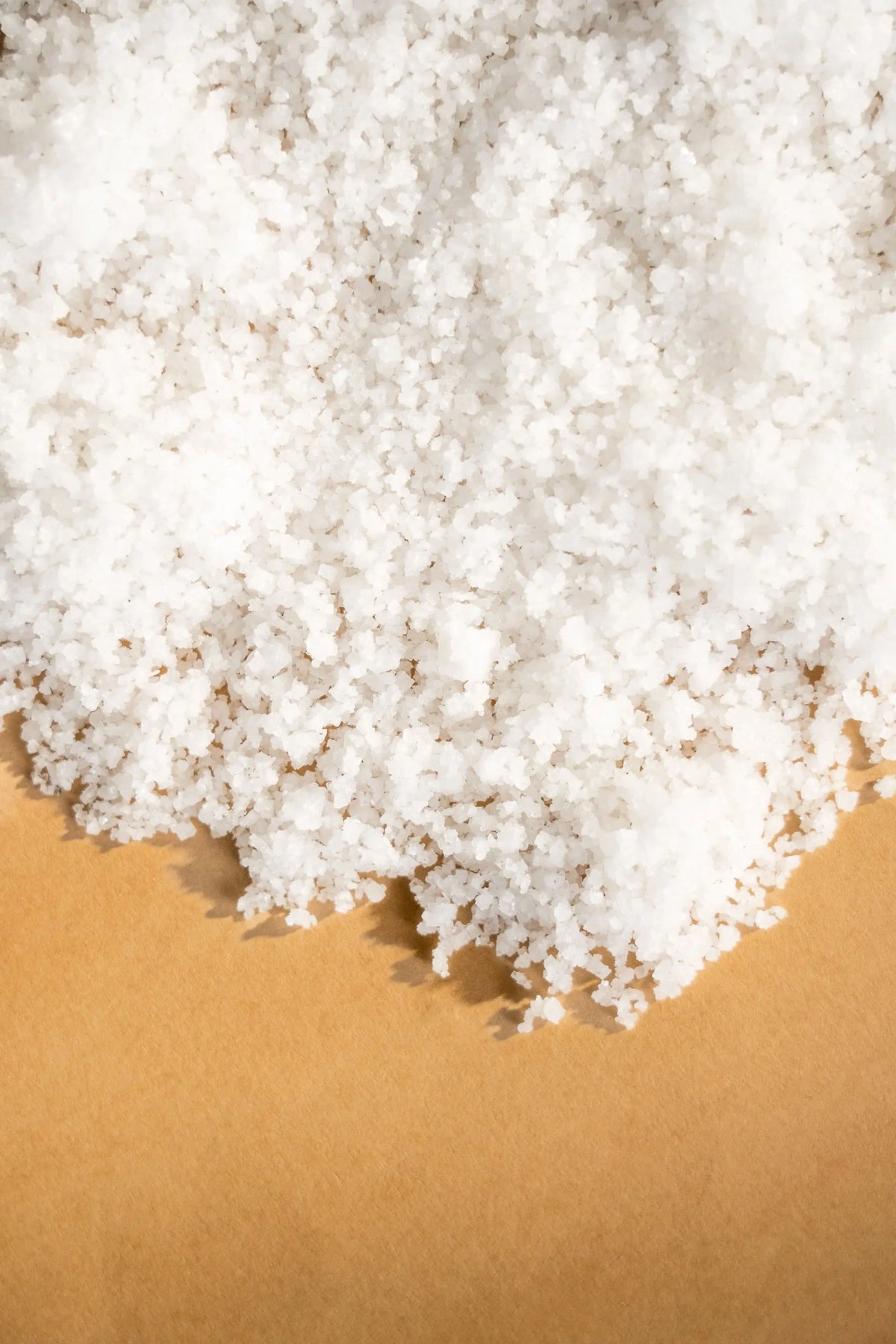 Close-up of white, coarse-grained artisanal spring salt crystals piled on a smooth, tan-colored surface.