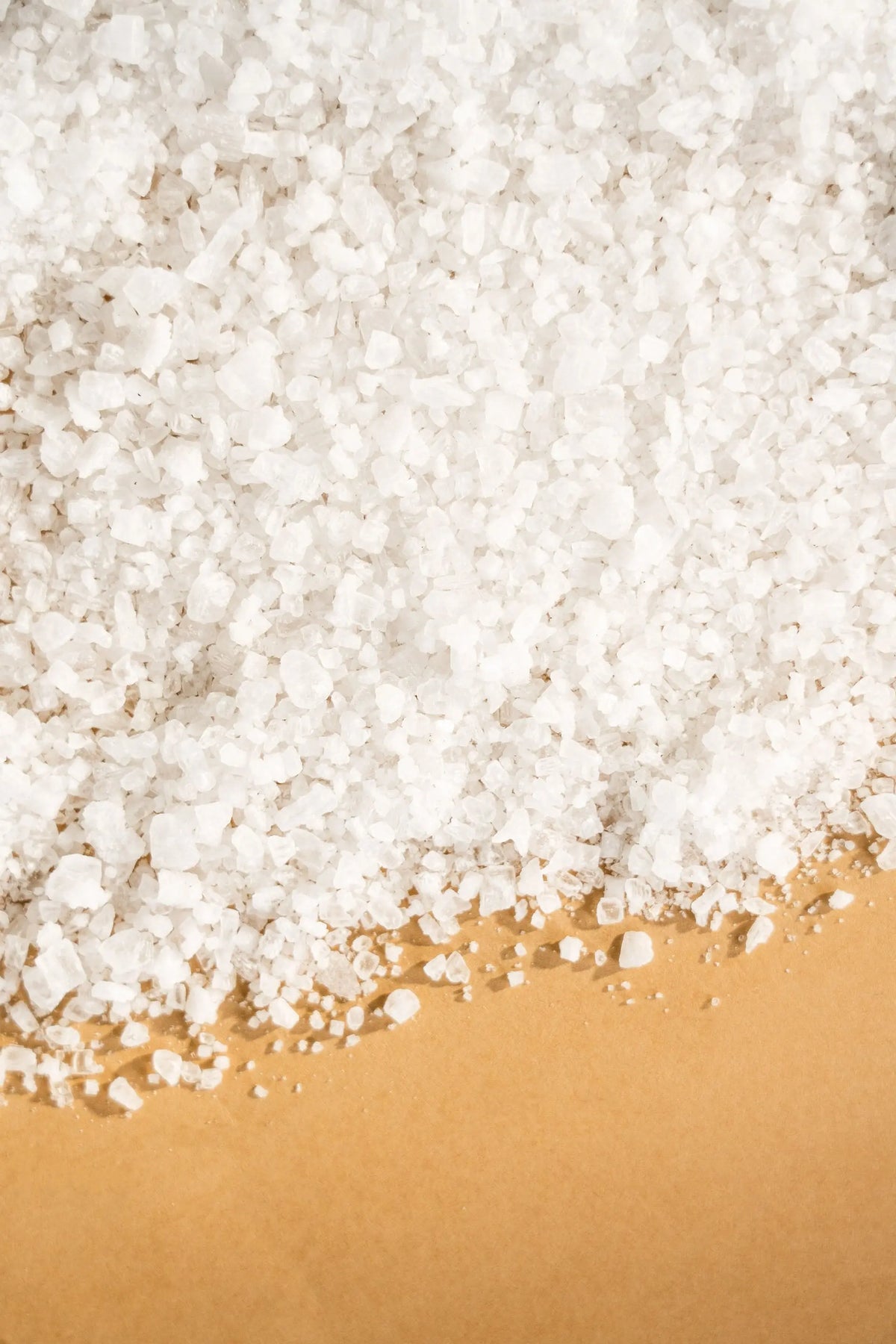 Coarse white natural salt crystals piled against a solid tan background, showing varied, crystalline shapes.