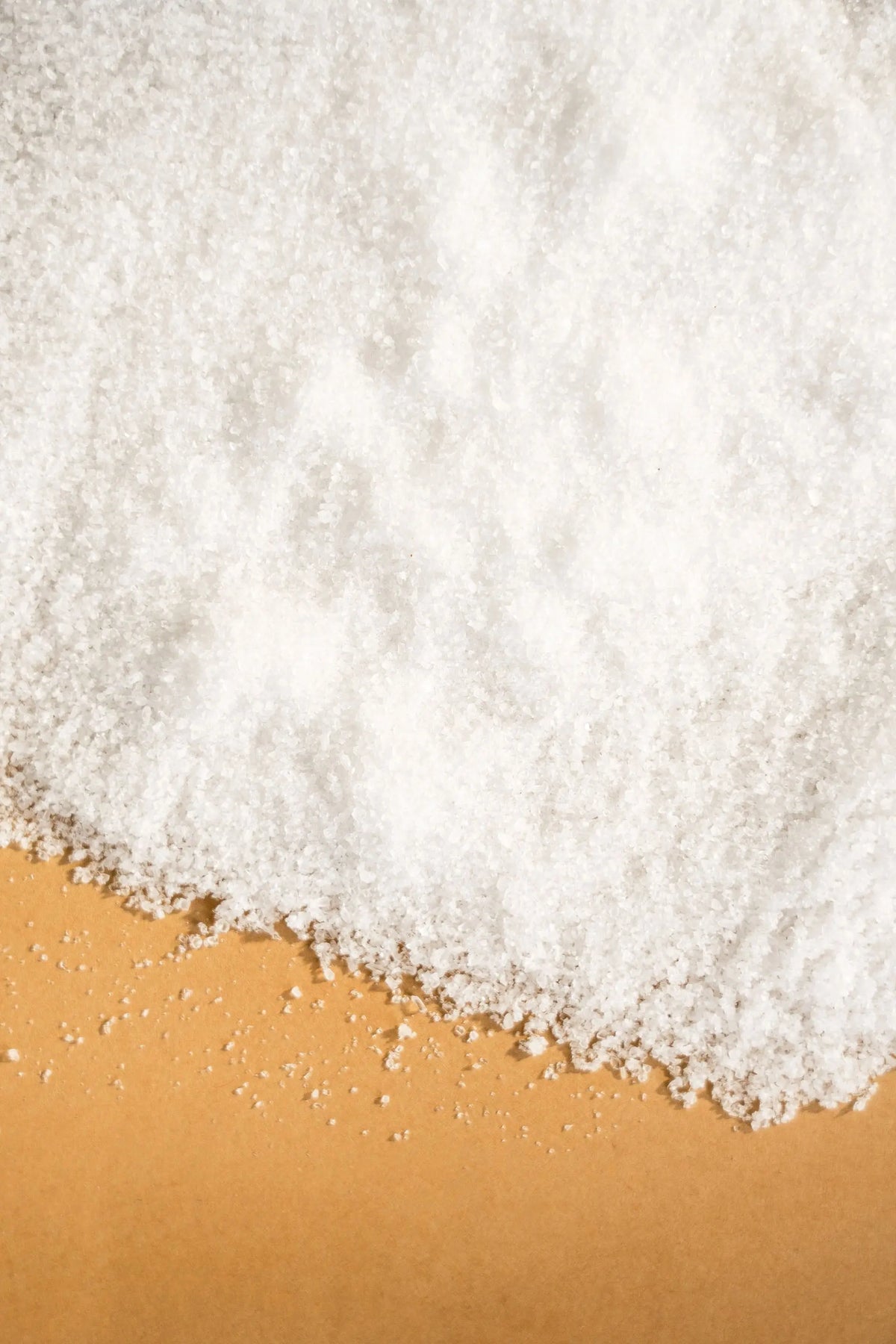 Pile of white, fine-grained natural spring salt scattered across a solid tan background.