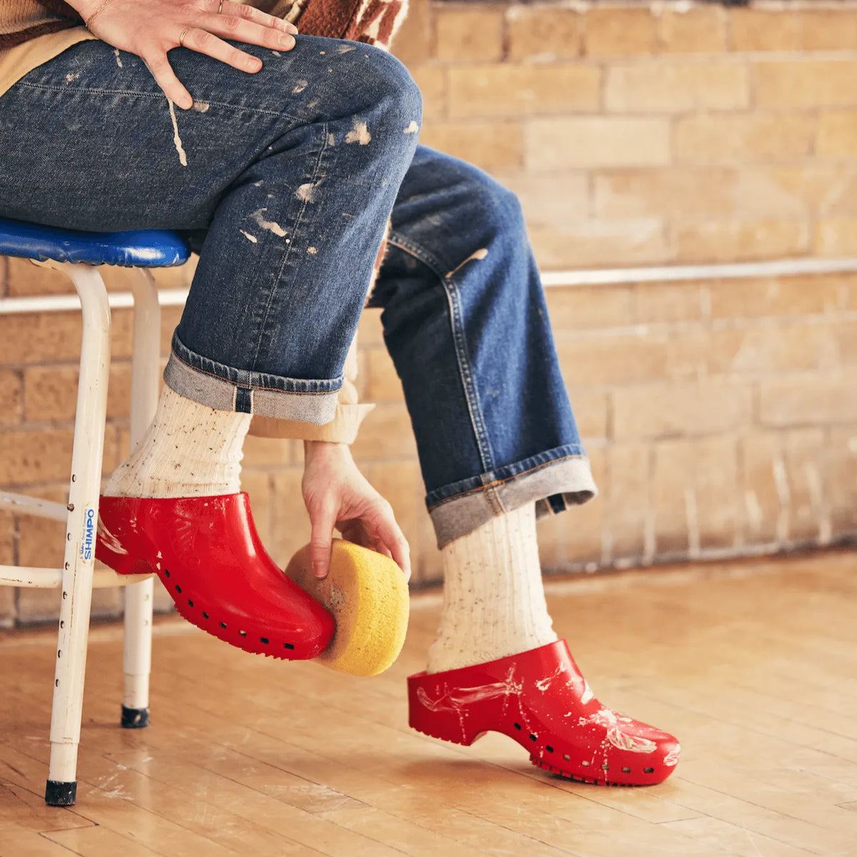 Person wearing vibrant red slip-on clogs with no holes, paired with speckled cream socks and paint-splattered jeans.