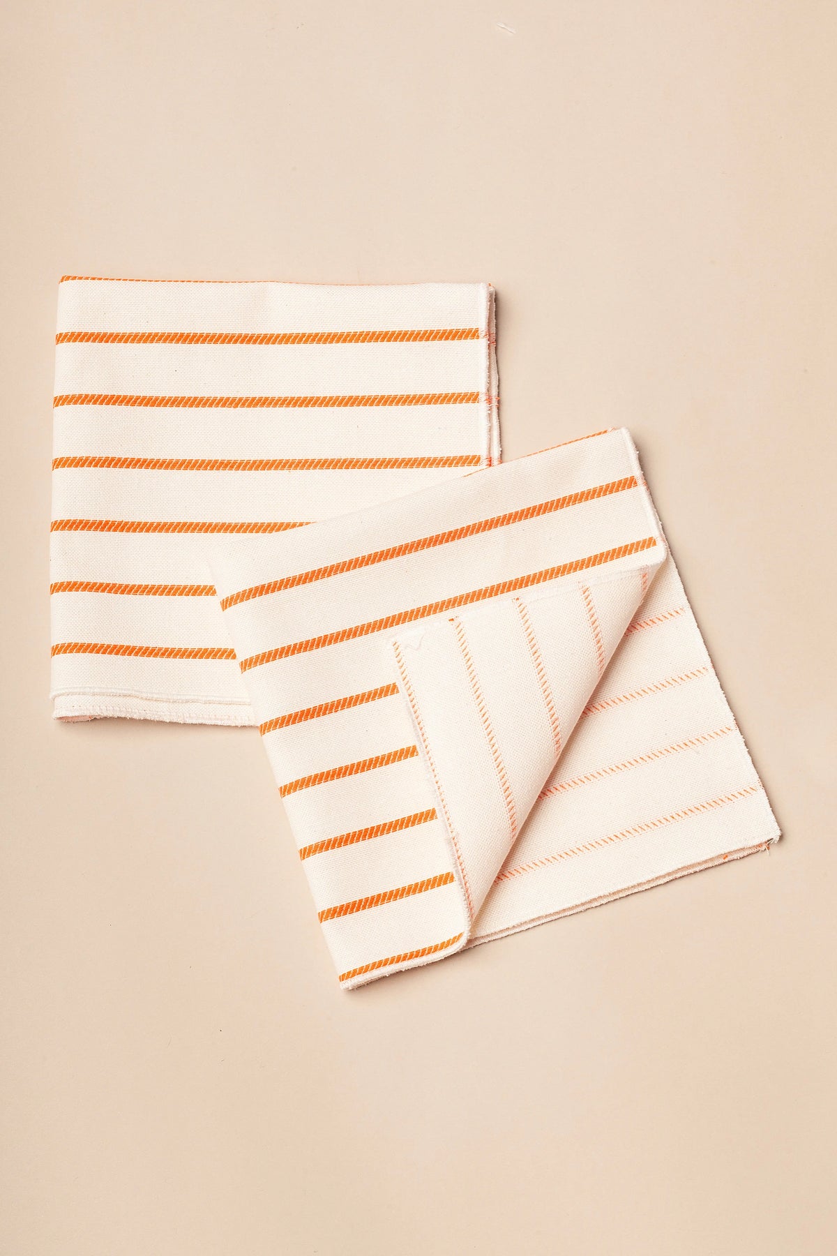 Pack de dos servilletas de tela color crema con rayas horizontales naranjas sobre fondo neutro.