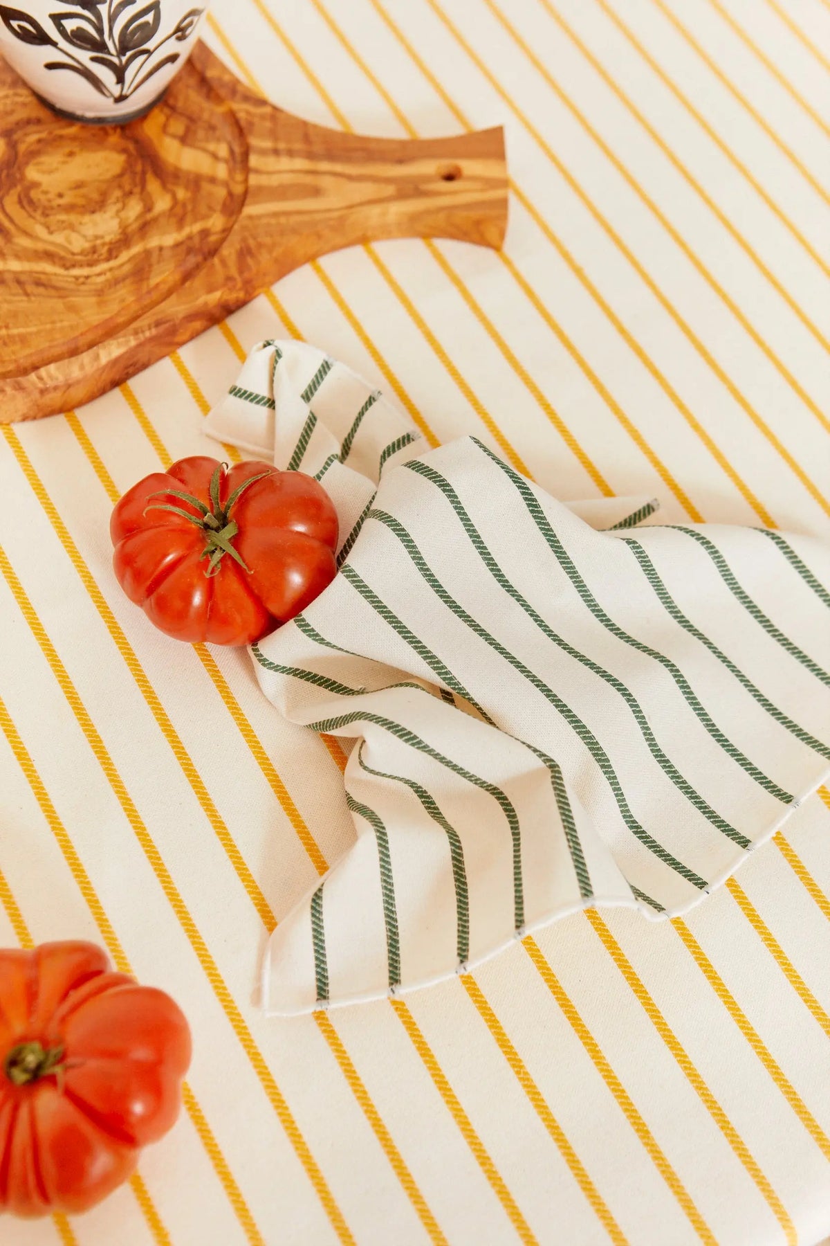 Pack de dos servilletas de tela con estampado de cadenas verdes sobre mantel de rayas amarillas y tomates frescos.