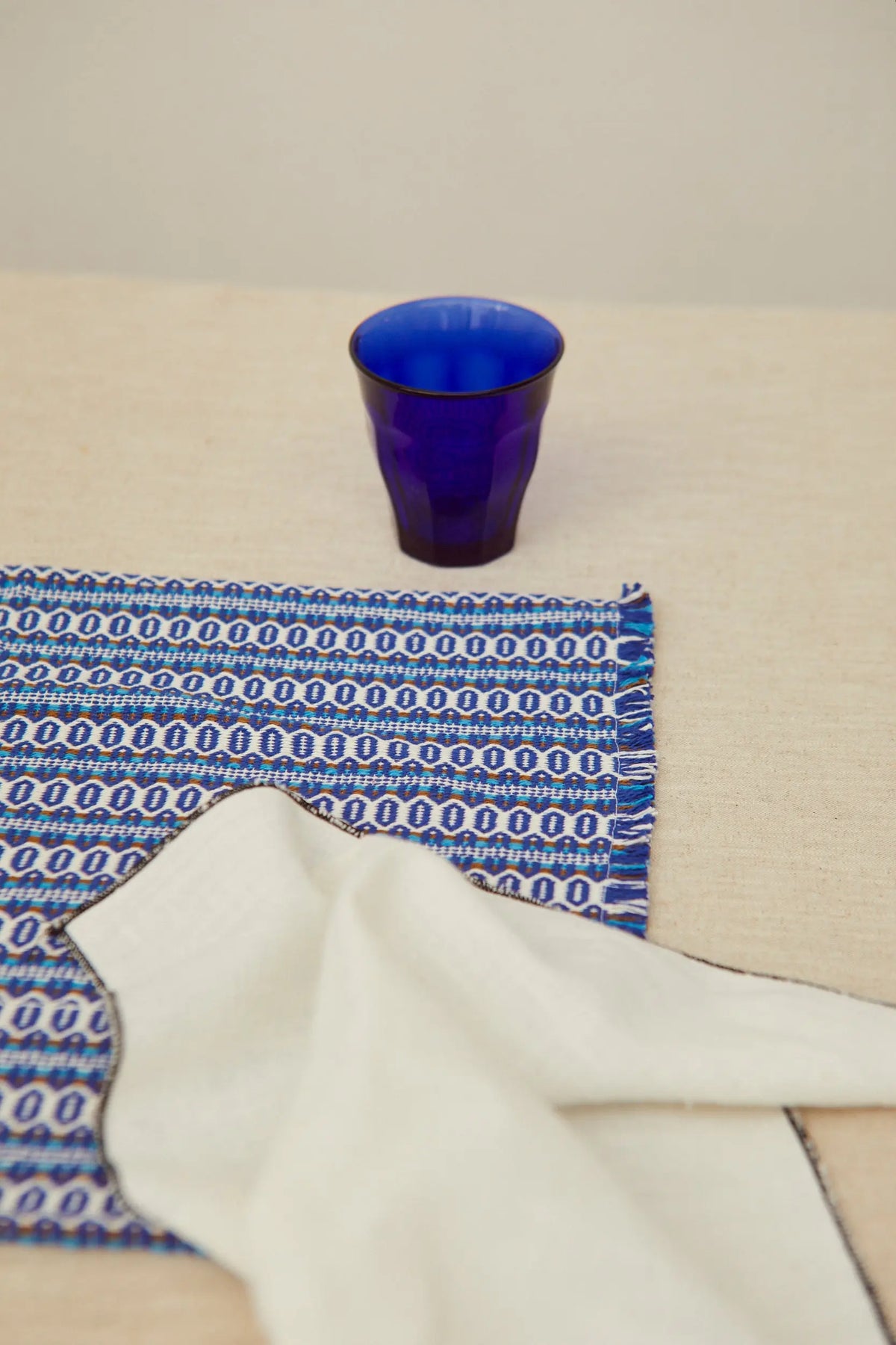 Pack de dos servilletas de lino blanco roto con bordes desflecados, sobre mantel azul estampado y vaso de cristal azul.