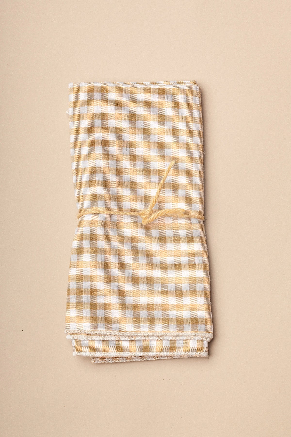 Pack de dos servilletas textiles con estampado de cuadros vichy beige, atadas con una cuerda fina sobre fondo neutro.