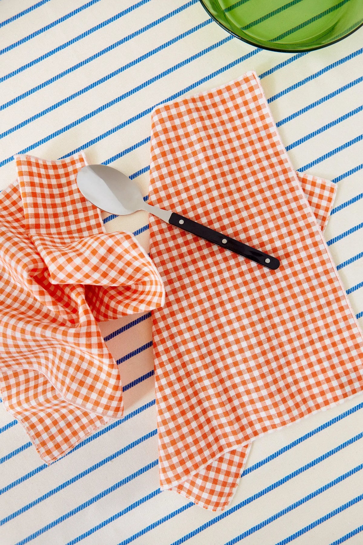 Pack de dos servilletas de tela con estampado de cuadros vichy naranjas sobre mantel con rayas azules.