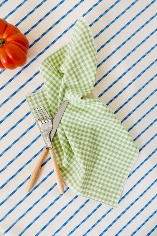Pack de dos servilletas de tela con estampado vichy verde sobre mantel de rayas azules, con cubiertos y un tomate.