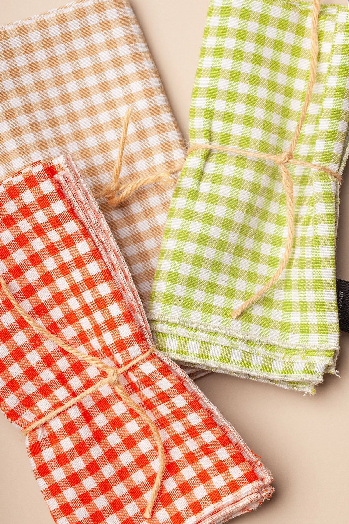Tres servilletas de tela con estampado vichy de cuadros pequeños en tonos verde, naranja y beige, atadas con cuerda.