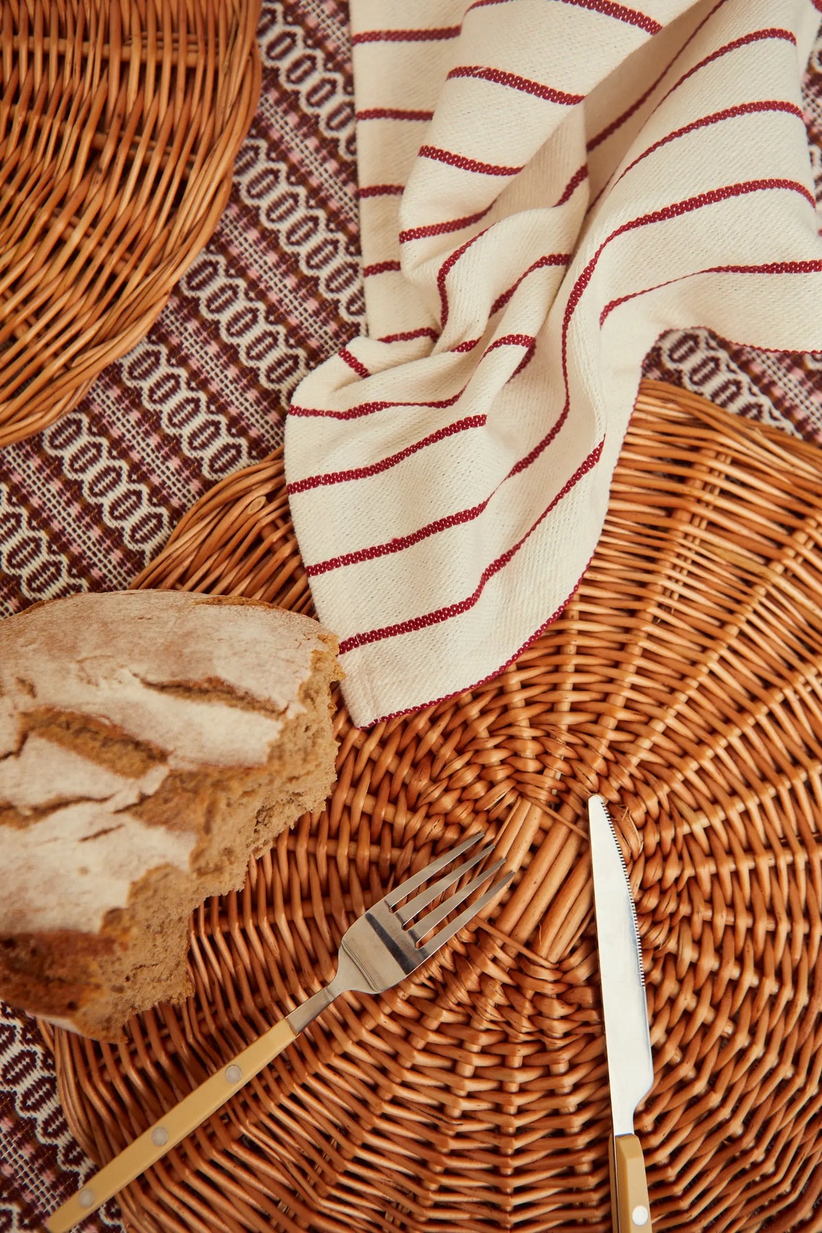Two cream-colored canvas napkins with thin burgundy stripes arranged on a woven wicker placemat with bread and cutlery.