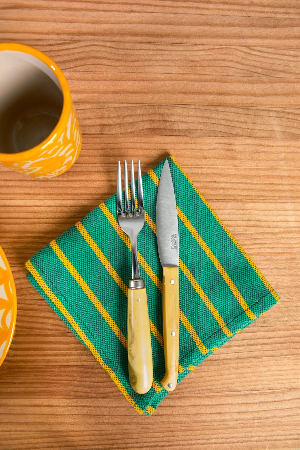 Servilleta de tela cuadrada con rayas verdes y amarillas sobre mesa de madera, con tenedor y cuchillo encima.