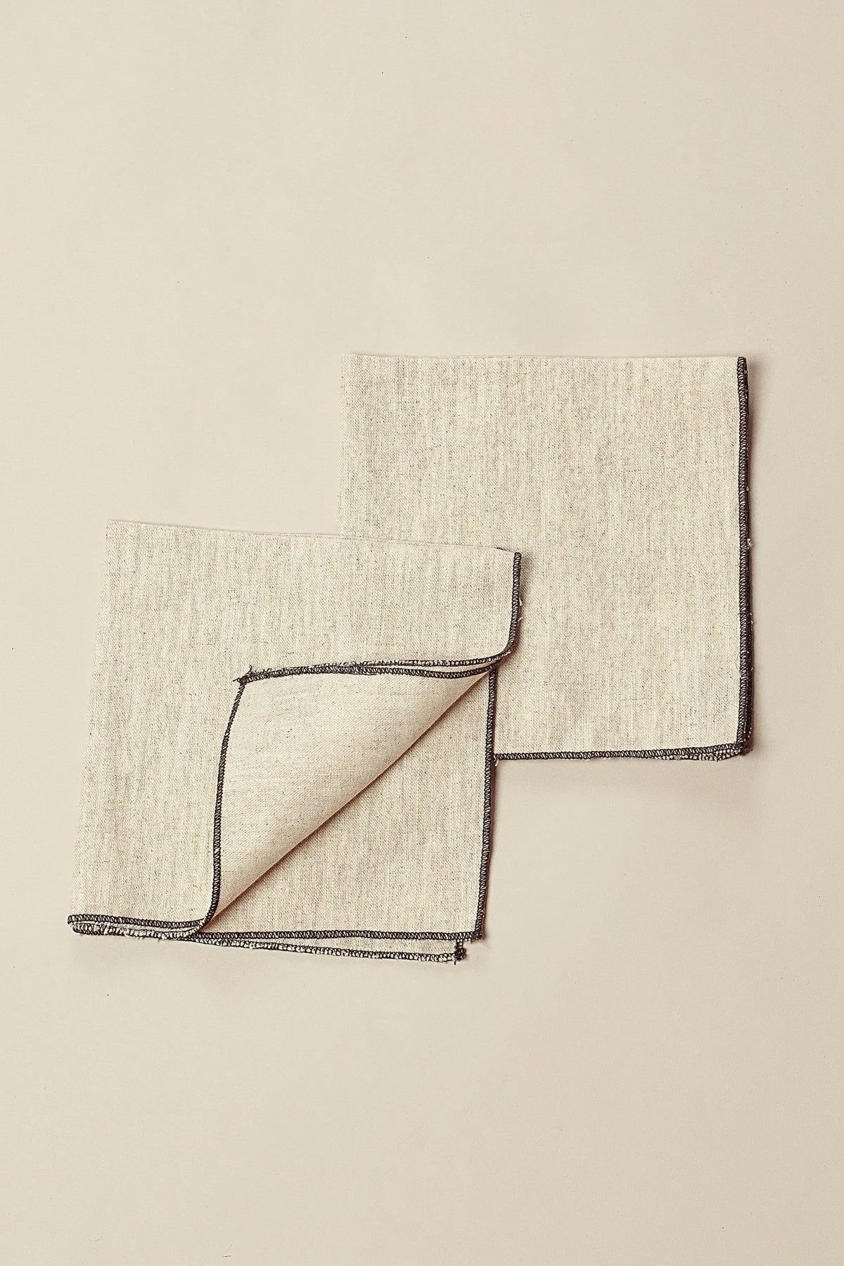 Pack de dos servilletas de lino beige, cuadradas, con acabado de costura en contraste oscuro en los bordes.