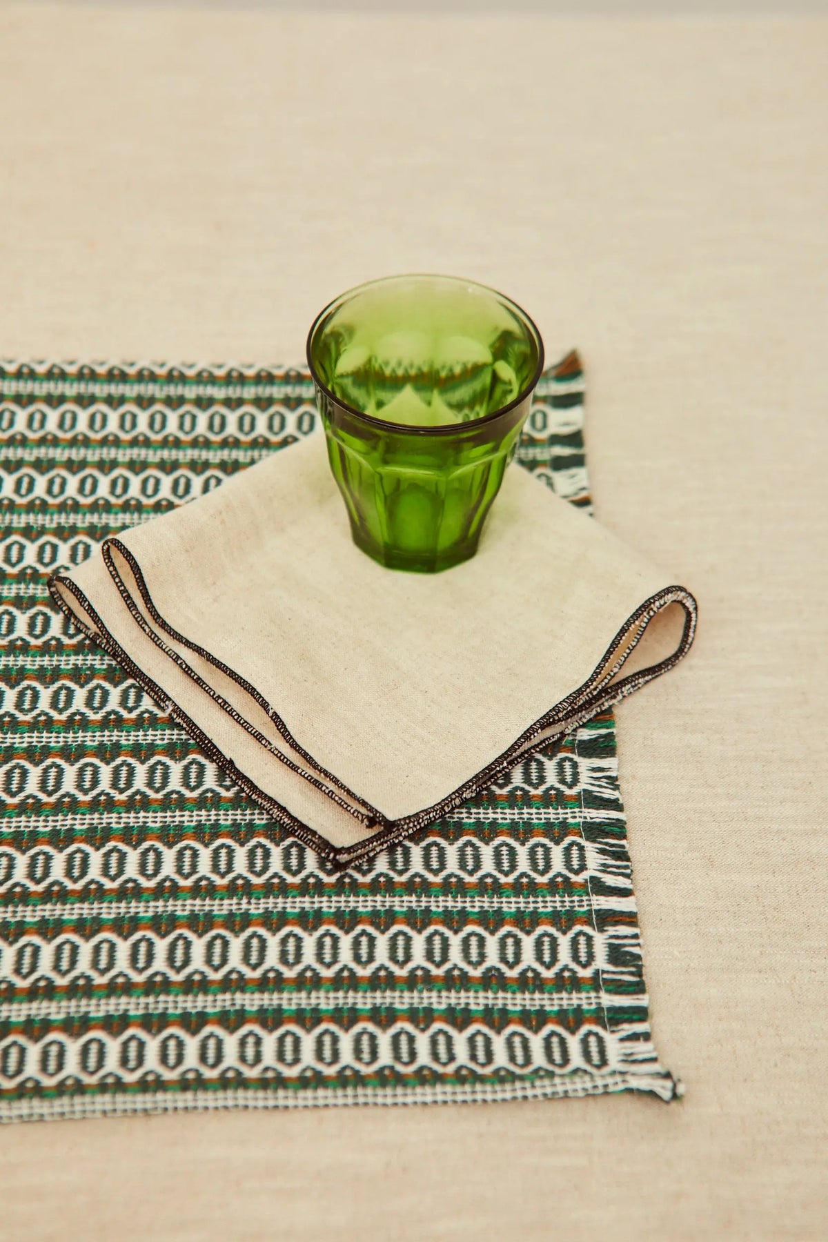 Servilleta de lino beige con borde oscuro sobre mantel estampado y un vaso de cristal verde.