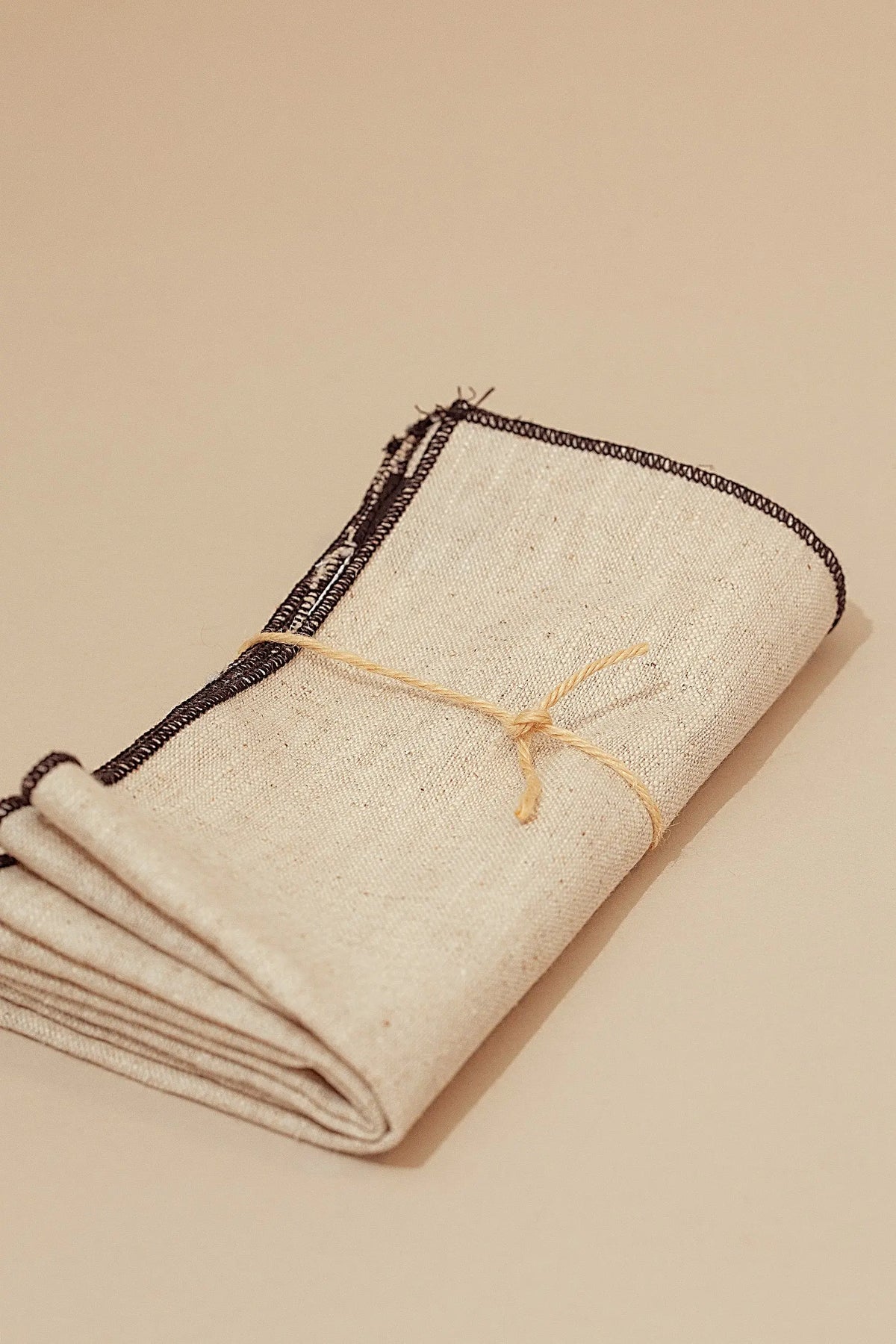 Dos servilletas de lino beige, dobladas, con bordes de costura negra y atadas con un cordel fino de fibra natural.