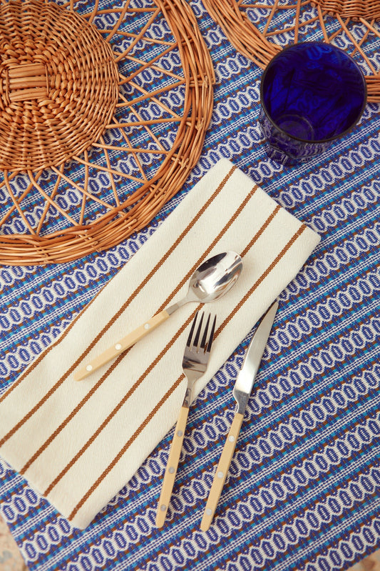 Off-white cotton napkin with caramel-colored stripes, topped with silverware on a blue patterned tablecloth.