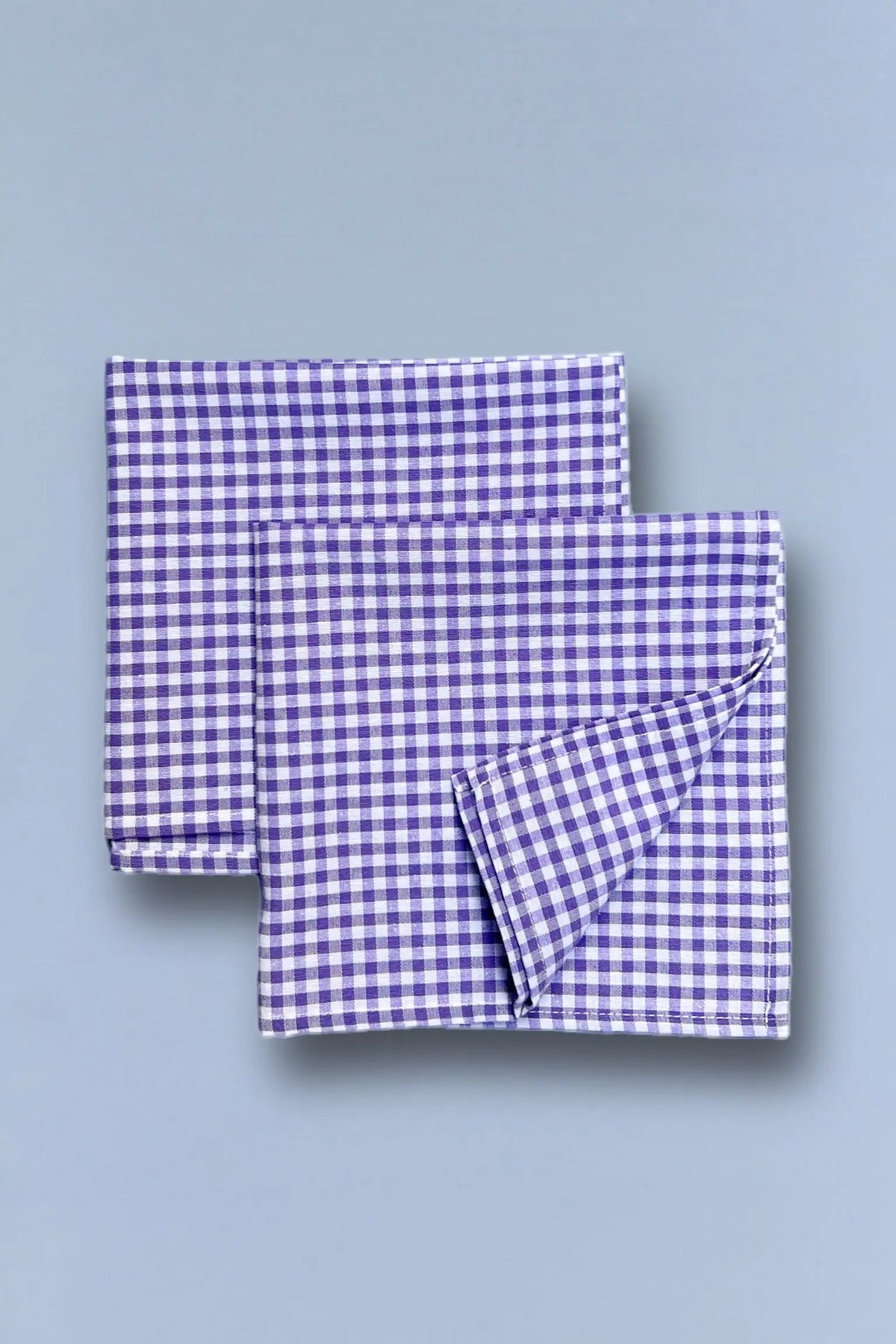 Two square napkins with a small-scale purple and white gingham check pattern, neatly folded on a light blue background.