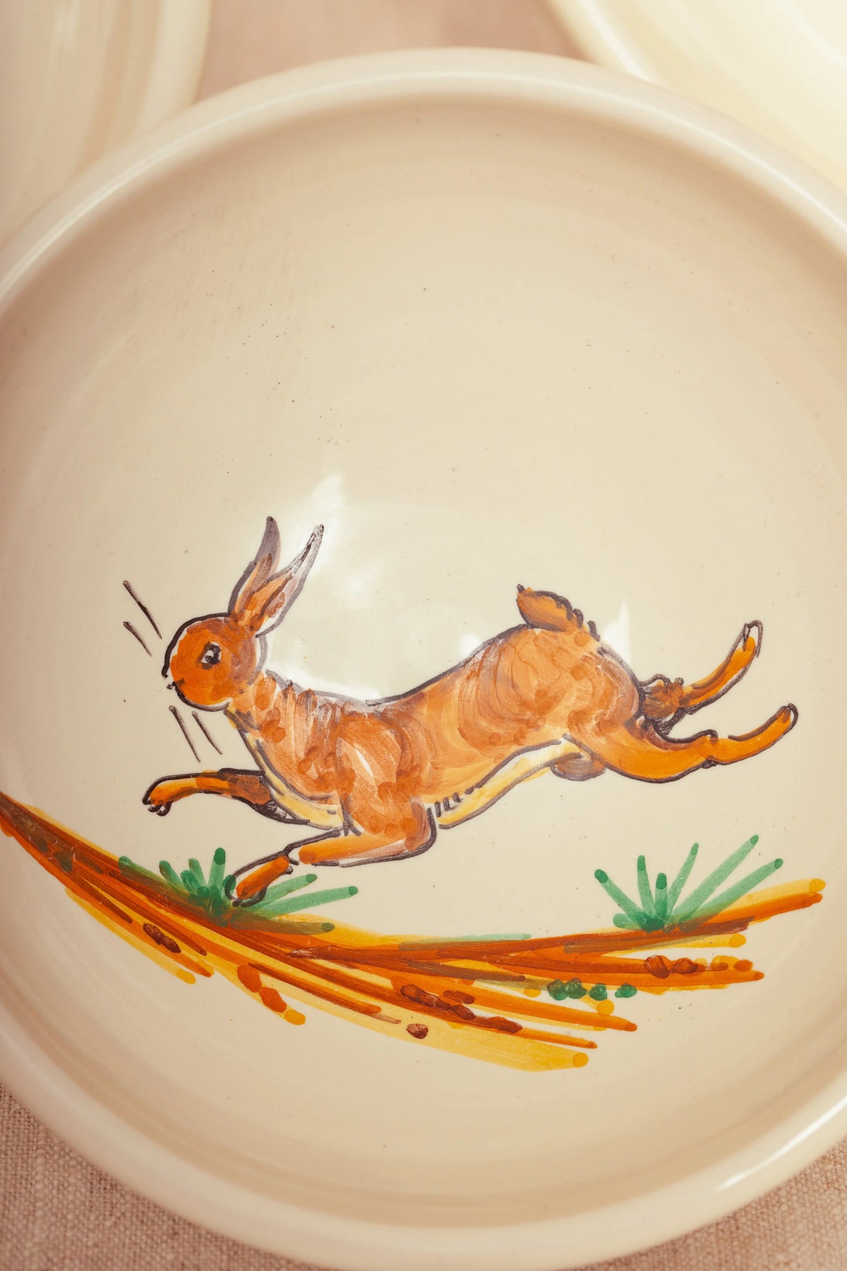 Cream ceramic bowl featuring a hand-painted brown running rabbit motif over a stylized landscape.