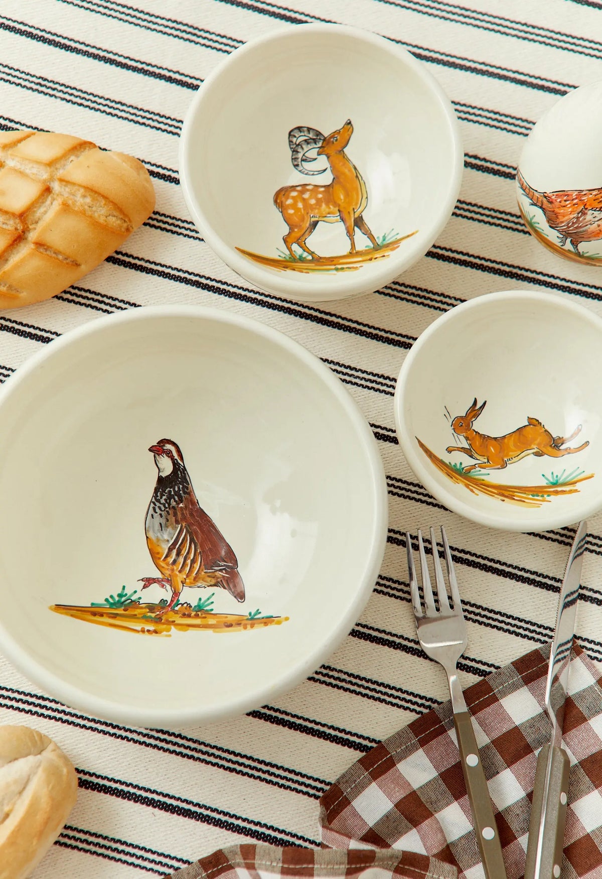 Set de cuencos de cerámica color crema con ilustraciones pintadas de animales del bosque sobre mantel a rayas.