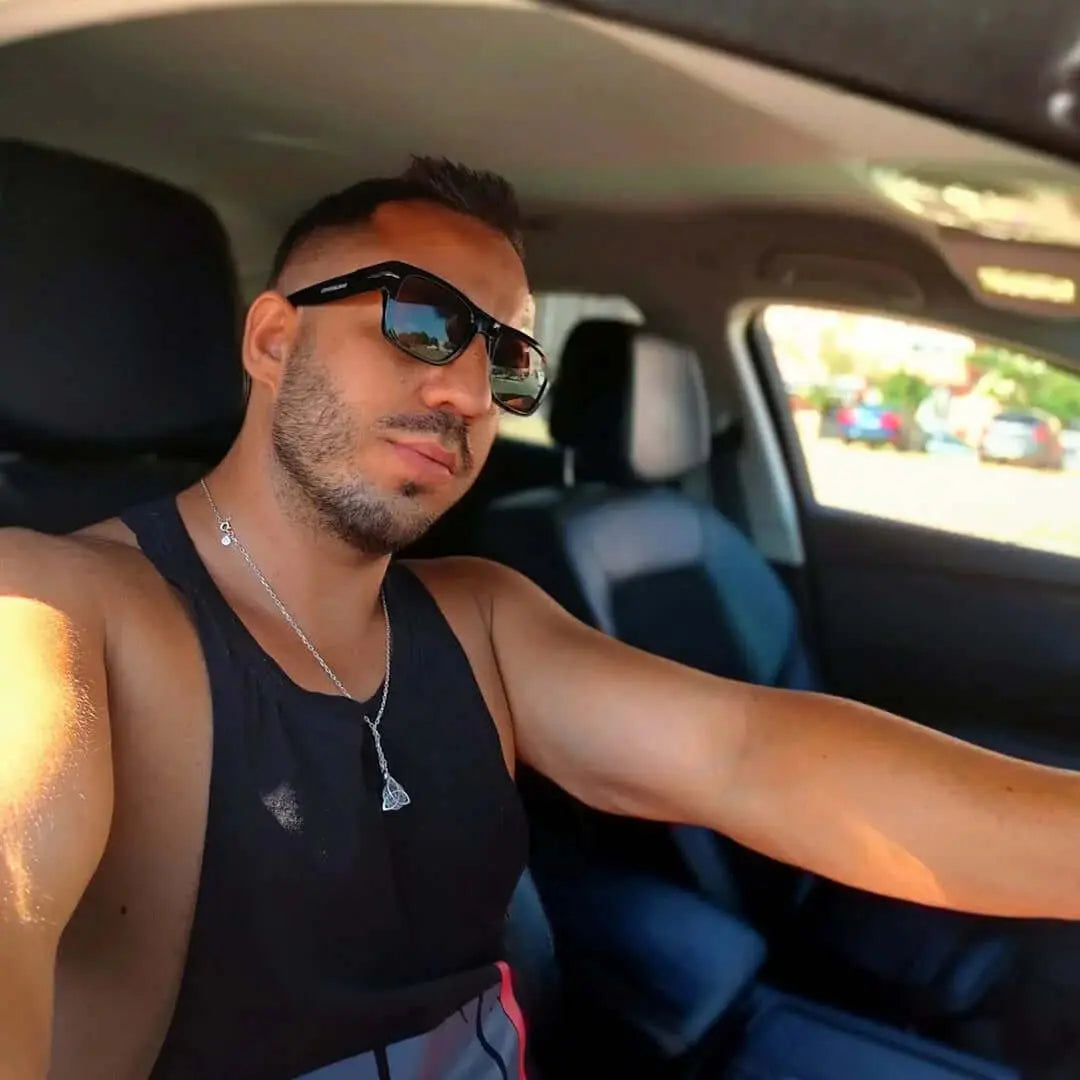 Hombre con gafas de sol retro de montura negra y lentes marrones conduciendo coche, estilo casual con camiseta negra.