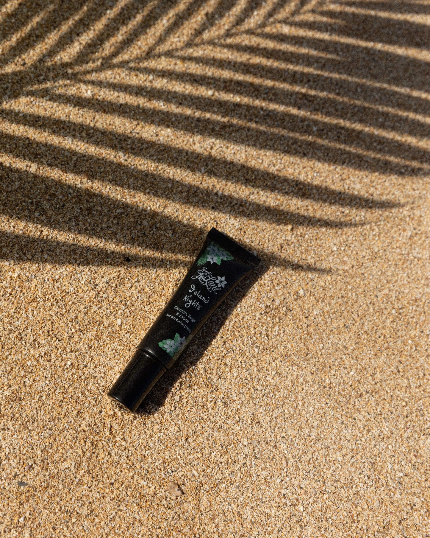 A black squeeze tube of Island Nights cream rests on golden sand under the shadow of a palm frond.